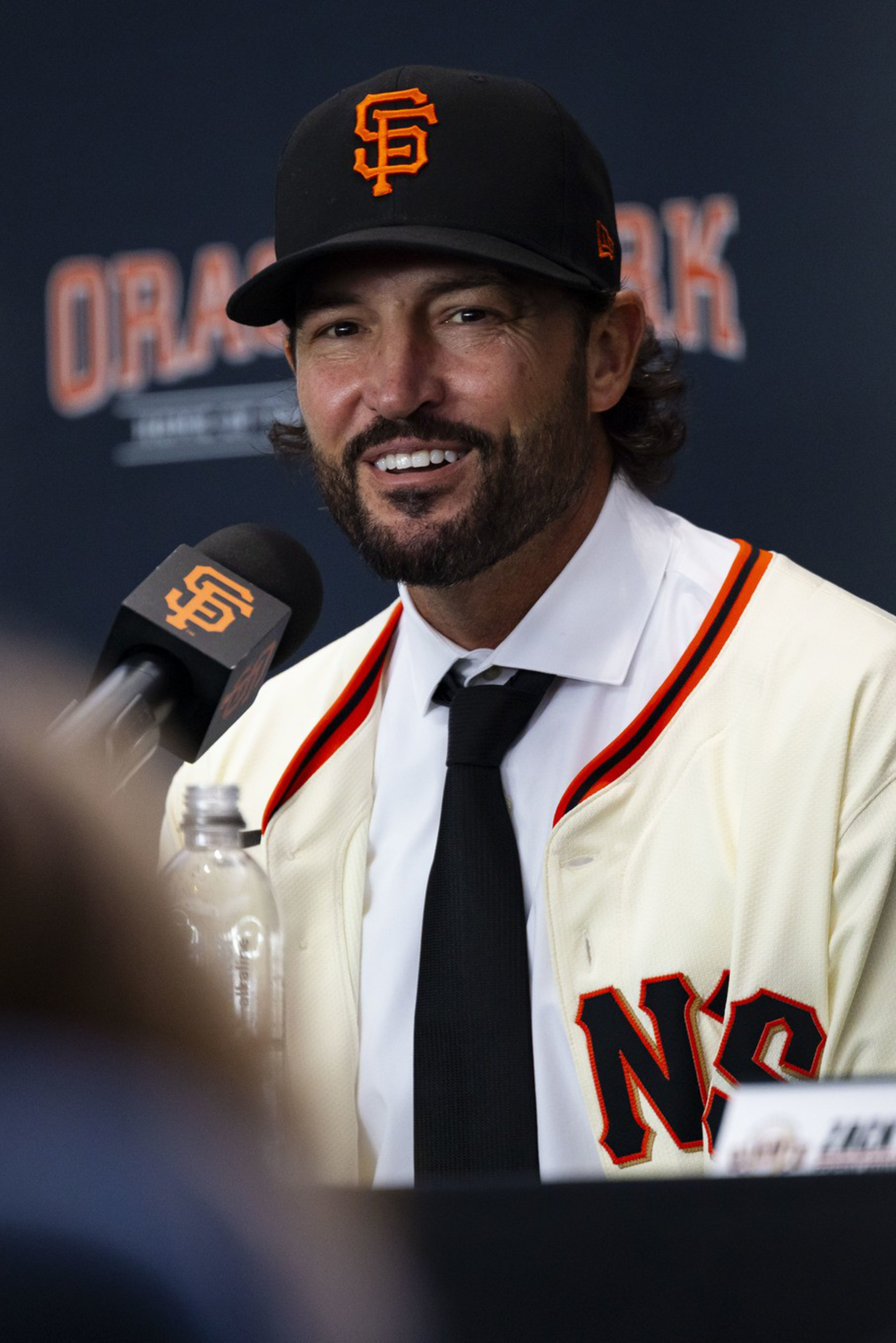 A man wearing a San Francisco Giants baseball cap and jersey smiles while speaking into a branded microphone at a press event.