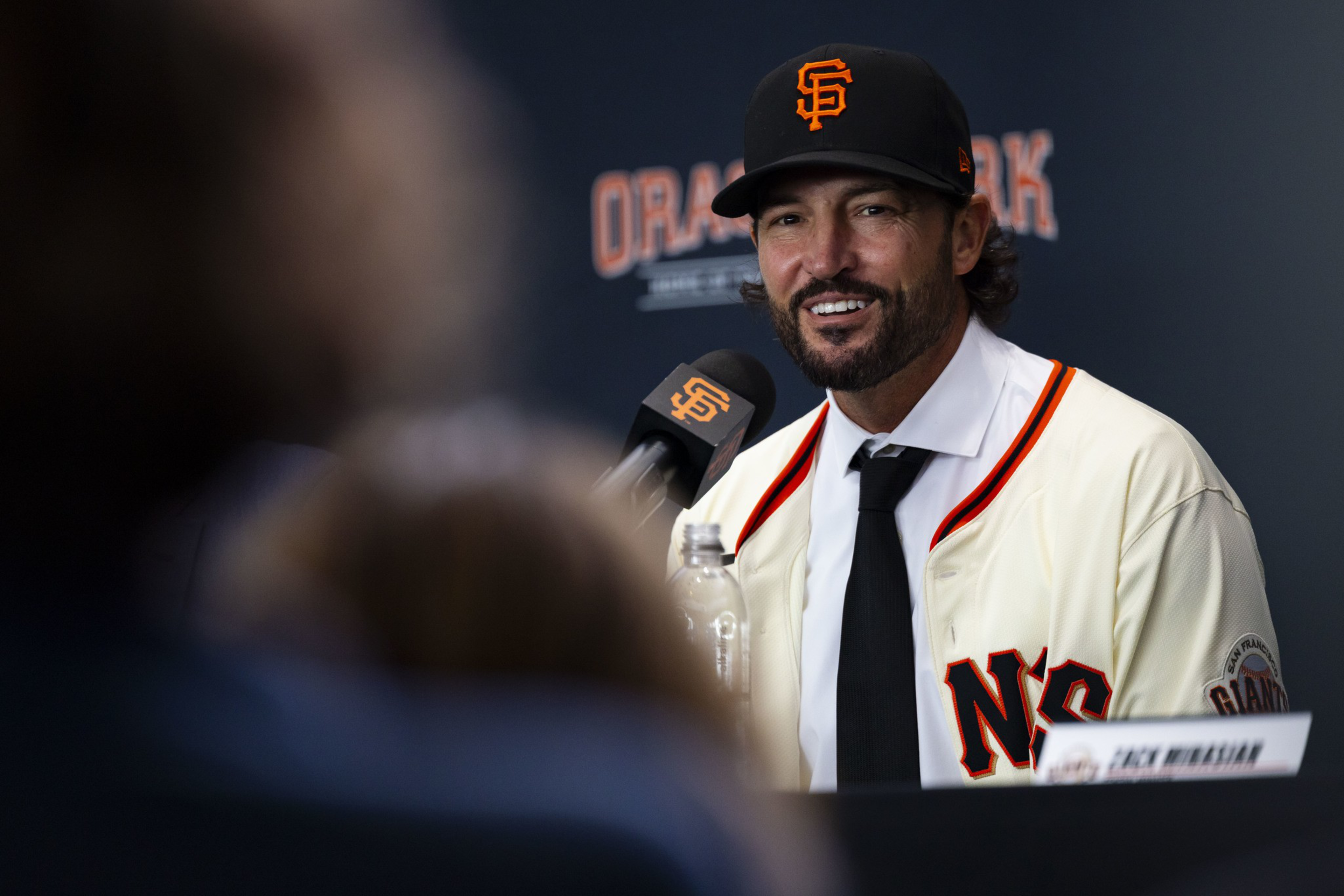 A man wearing a San Francisco Giants baseball cap and jersey smiles while speaking into a branded microphone at a press event.