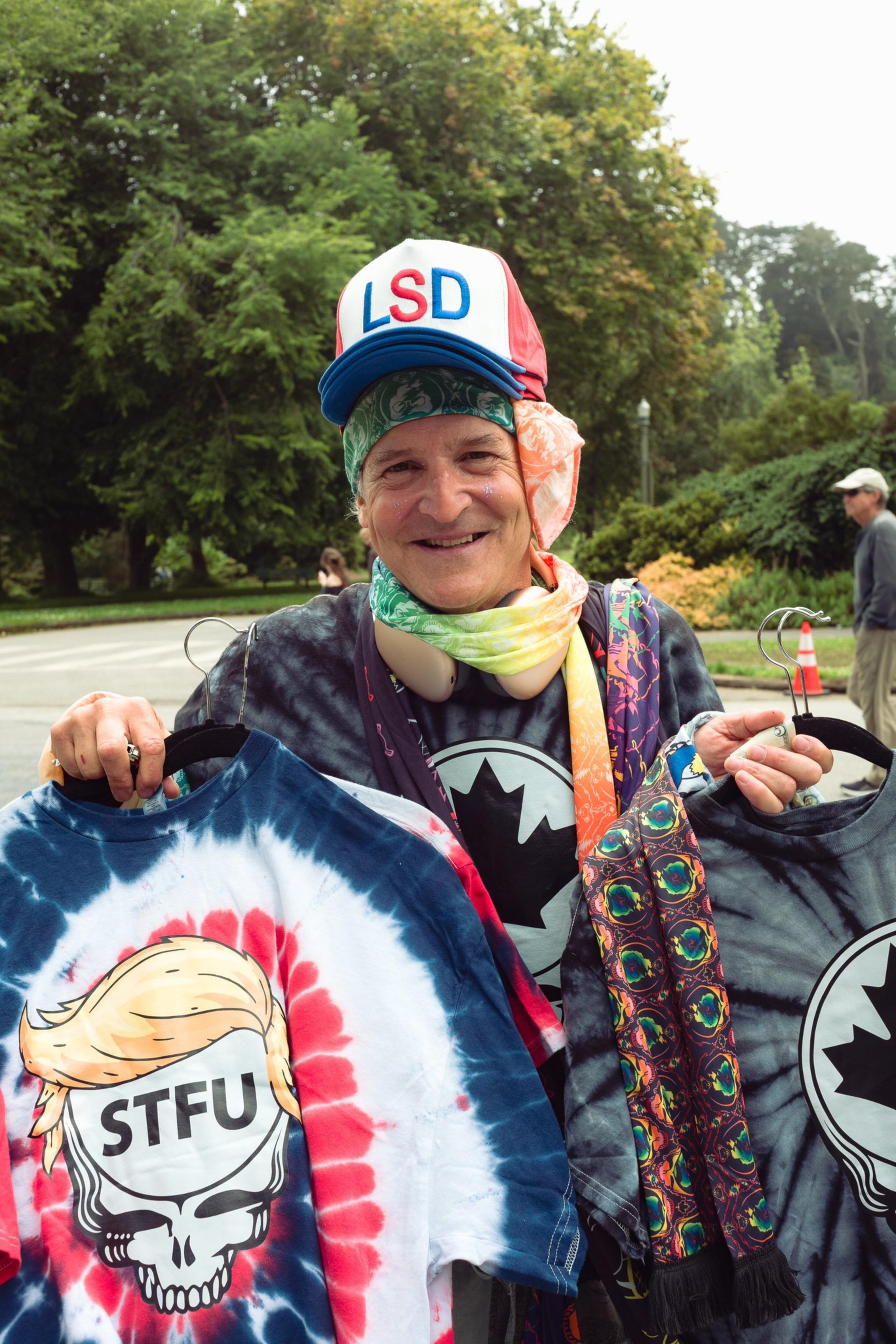 A smiling person wearing a colorful bandana and an LSD cap holds two tie-dye shirts, one with "STFU" over a skull with blonde hair.