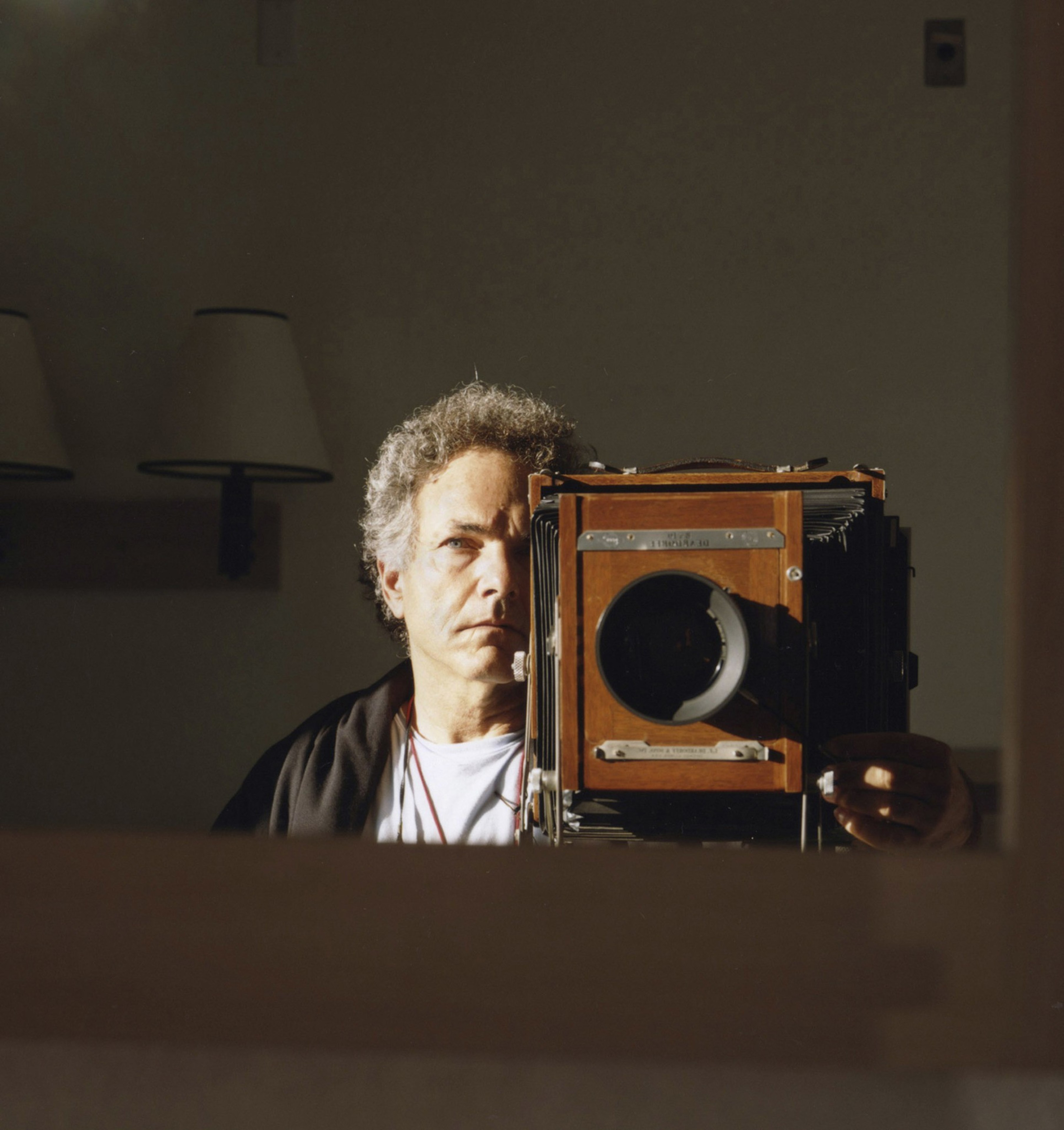 A person with curly gray hair is taking a selfie in a mirror using a large wooden camera. Two wall lamps are visible in the background.