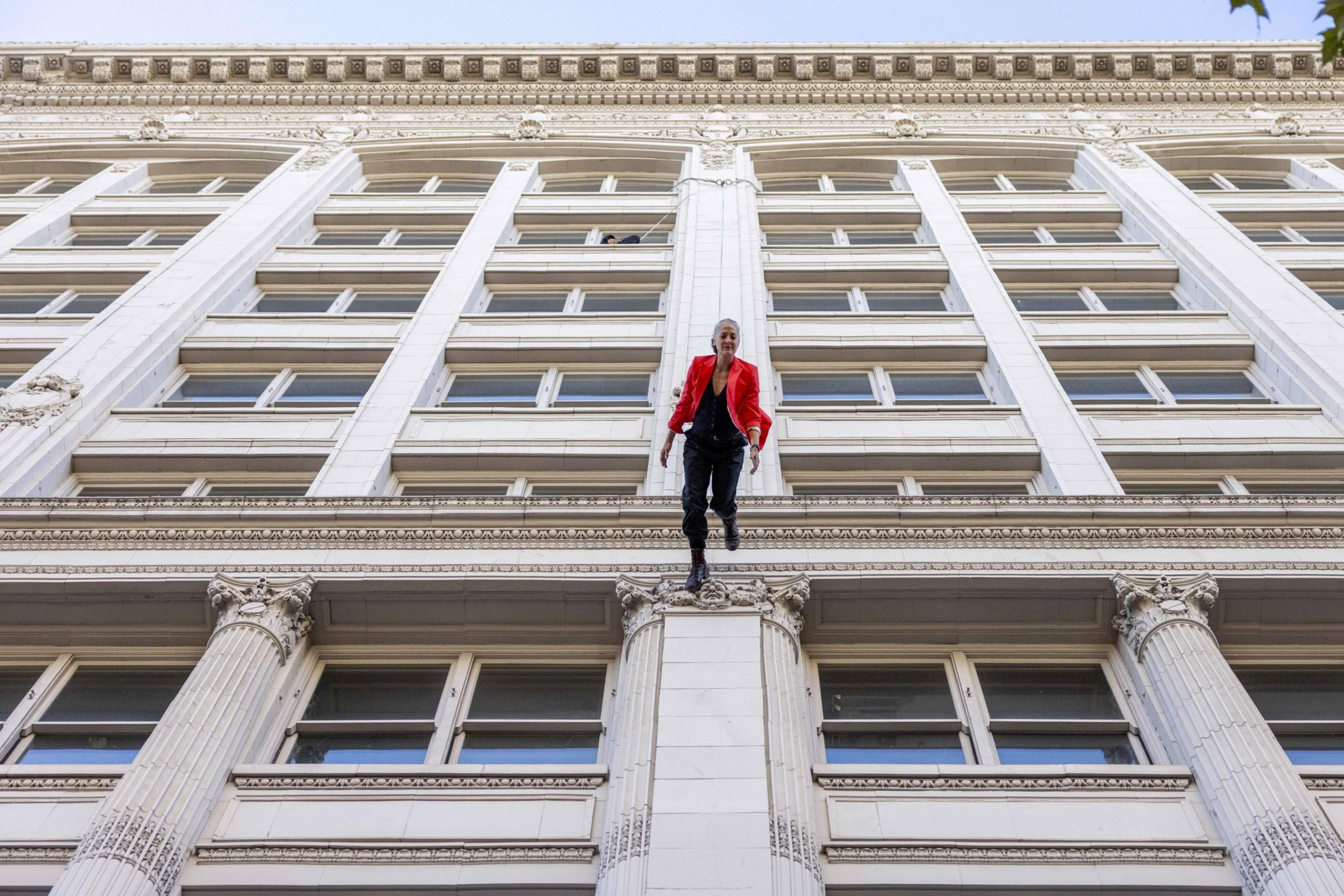 A person in a red jacket is dangling from the edge of a tall, ornate building with large windows and decorative columns, set against a clear blue sky.