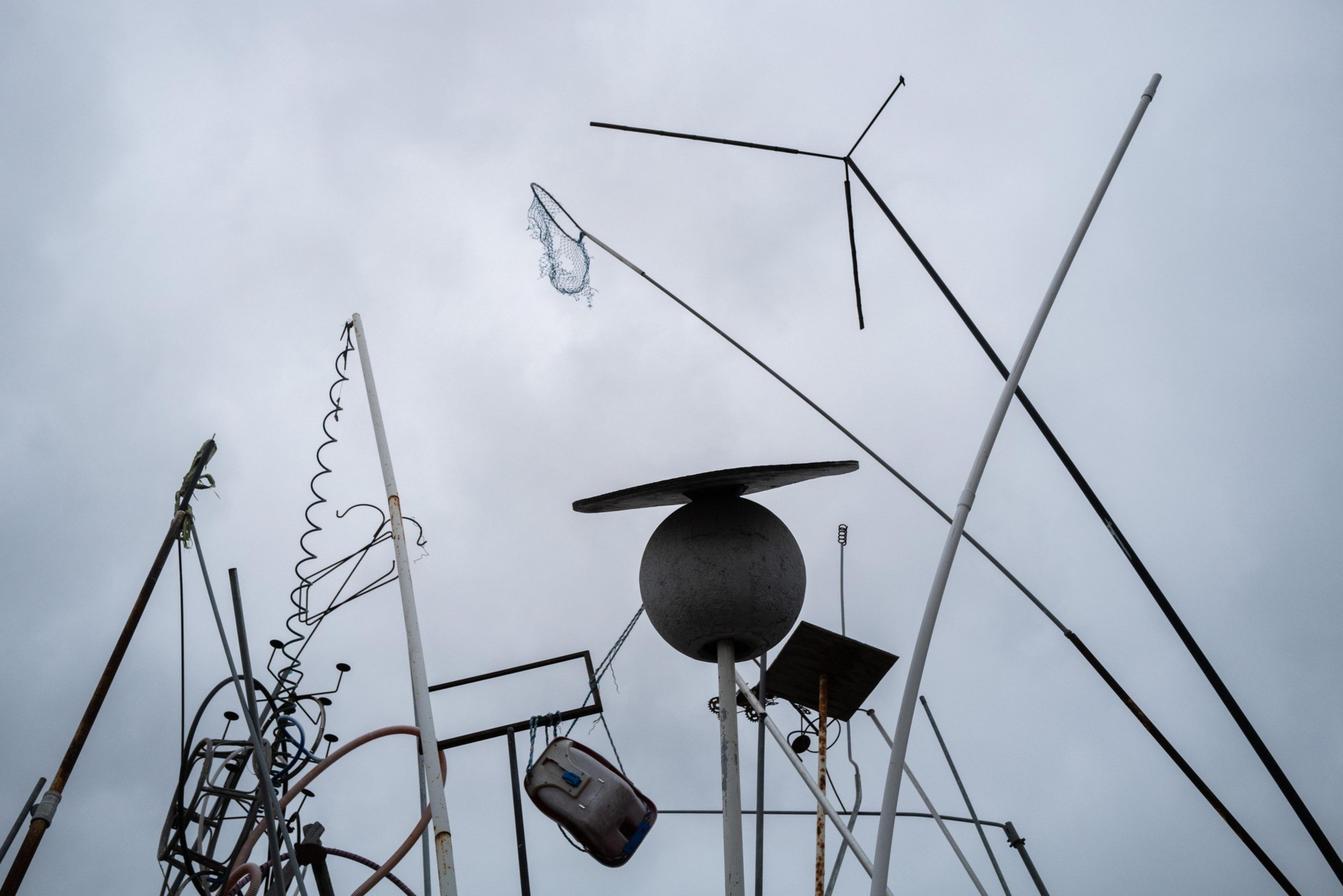 A collection of metal poles and rods of various shapes and sizes point skyward against a cloudy sky, with some twisted and others holding objects.