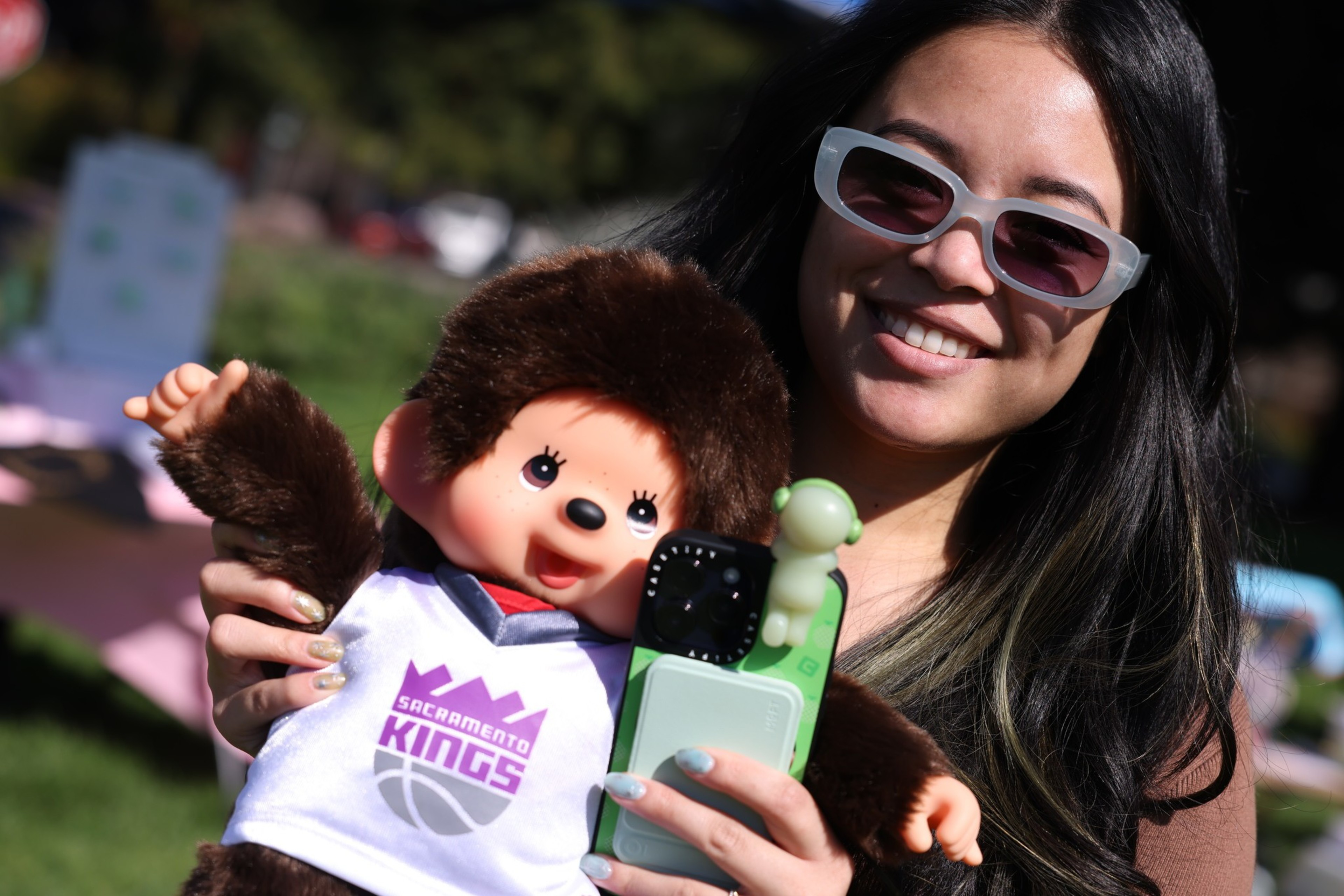 A smiling person in sunglasses holds a plush toy dressed in a Sacramento Kings jersey, along with a phone in a green case featuring a small green figure.