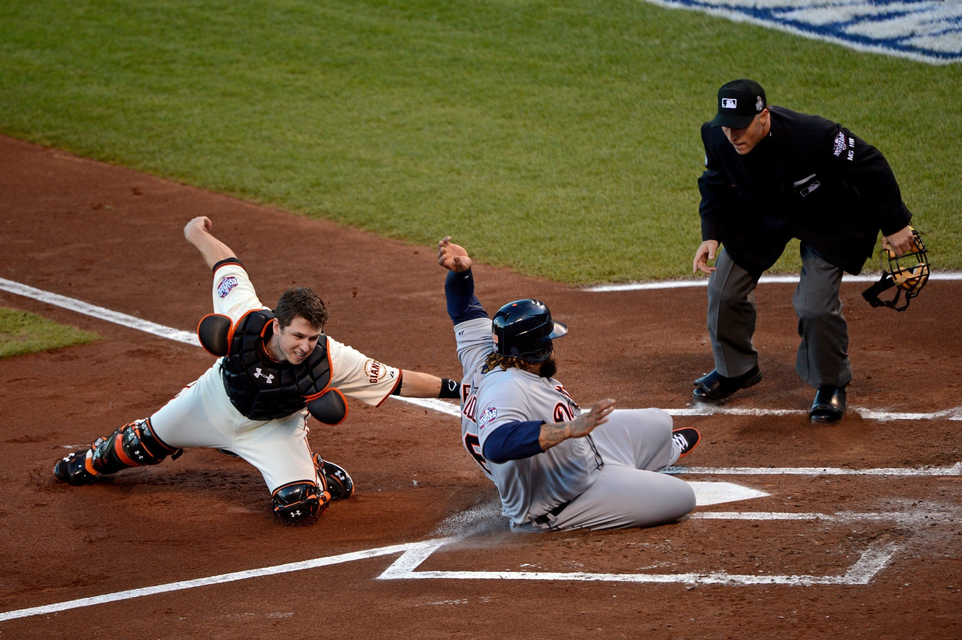 A baseball catcher in white gear tags a sliding runner in gray near home plate as an umpire watches closely, ready to make a call.
