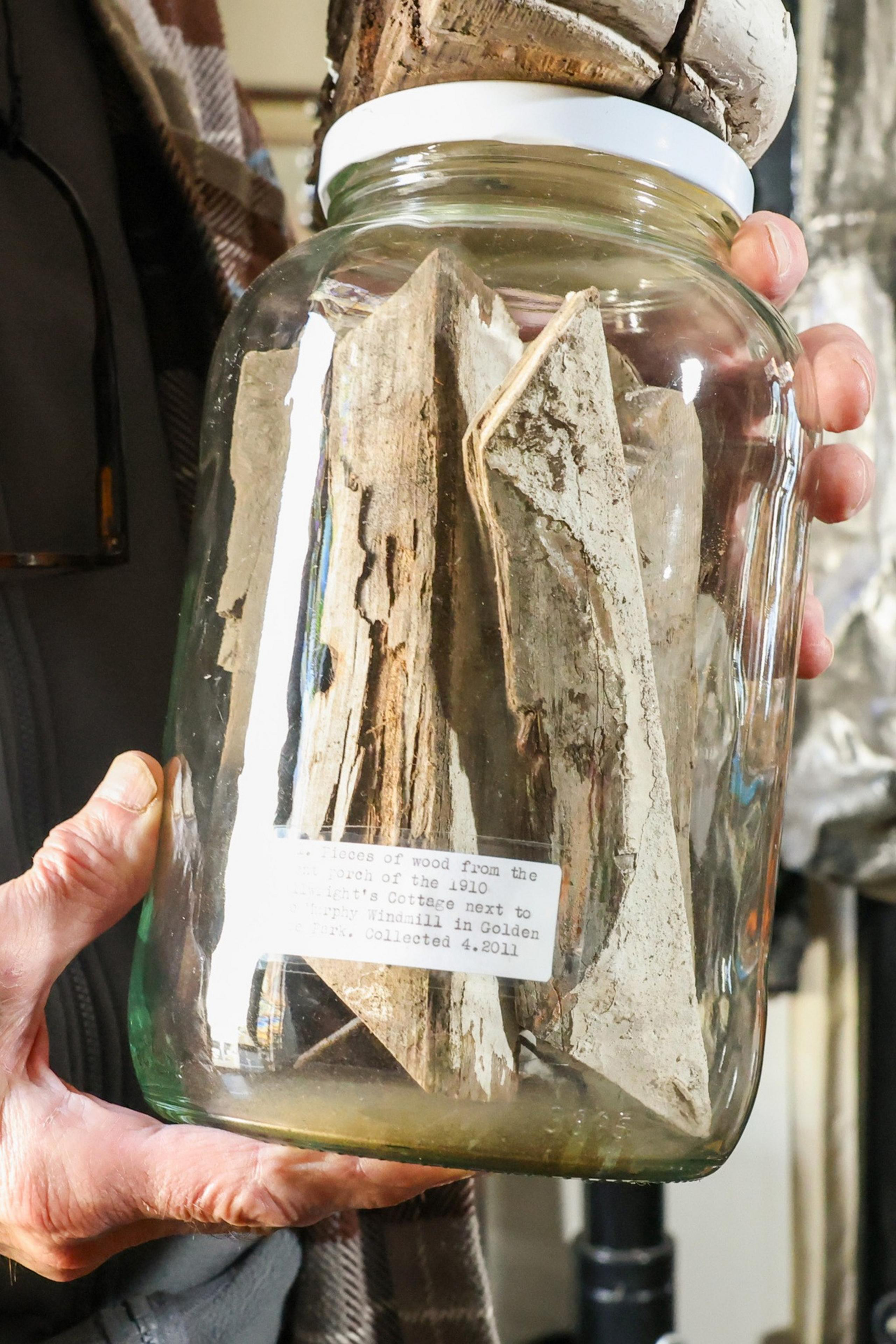 A hand holds a glass jar with pieces of aged wood inside. A label on the jar describes the wood's historical significance and collection date.