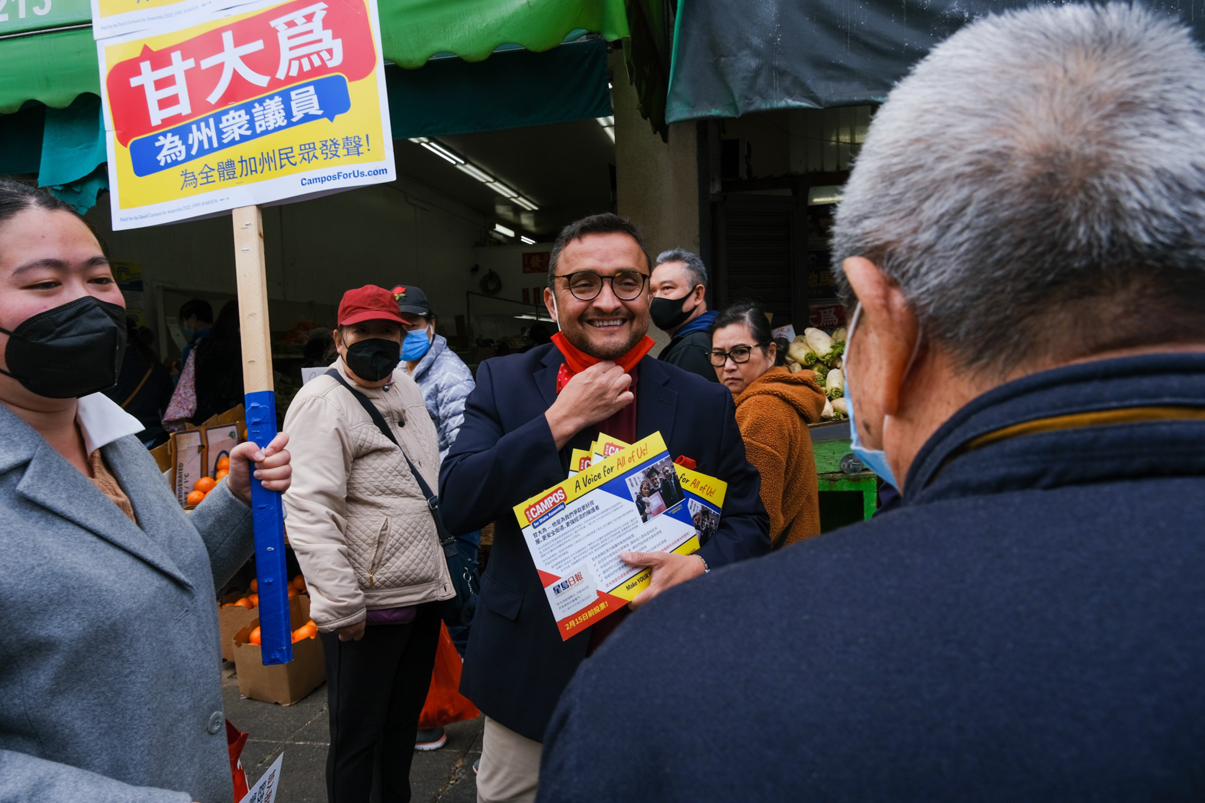 A man wearing glasses and a suit holds campaign flyers and smiles while adjusting his red face mask, surrounded by people, one holding a sign with Chinese text.