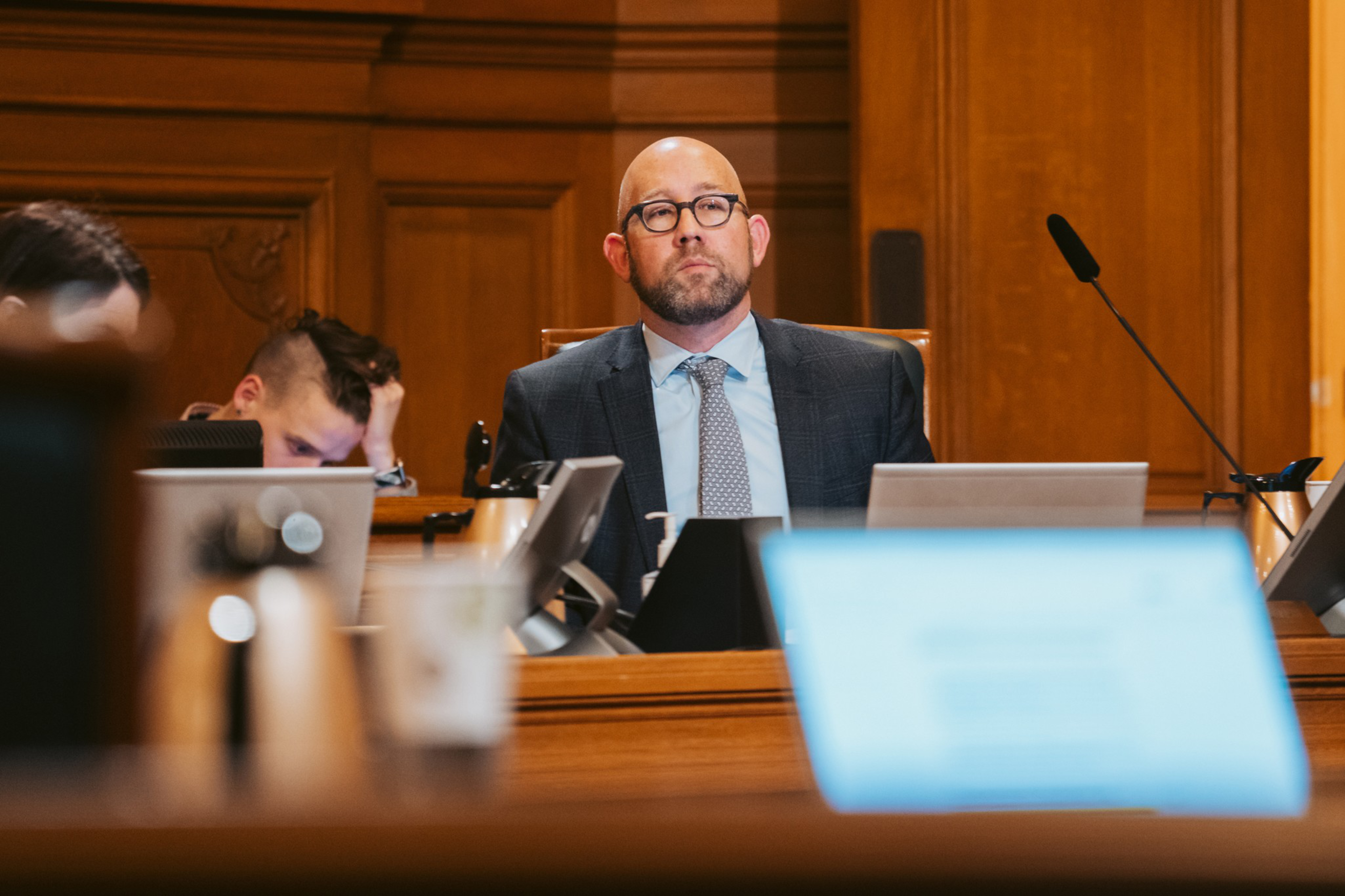 A man in a suit and glasses sits in a formal setting, surrounded by laptops and papers. Another person appears focused on a device beside him.