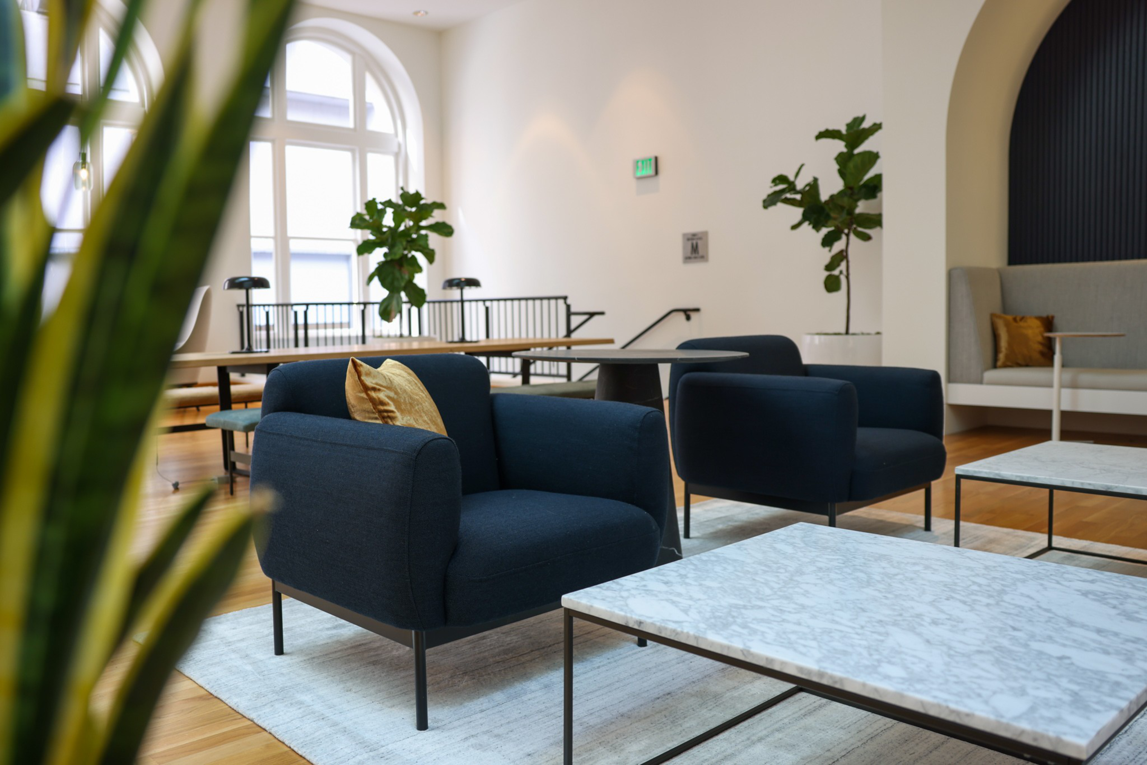 Two navy blue armchairs with yellow cushions sit around white marble coffee tables on a rug in a bright room with large arched windows and potted plants.