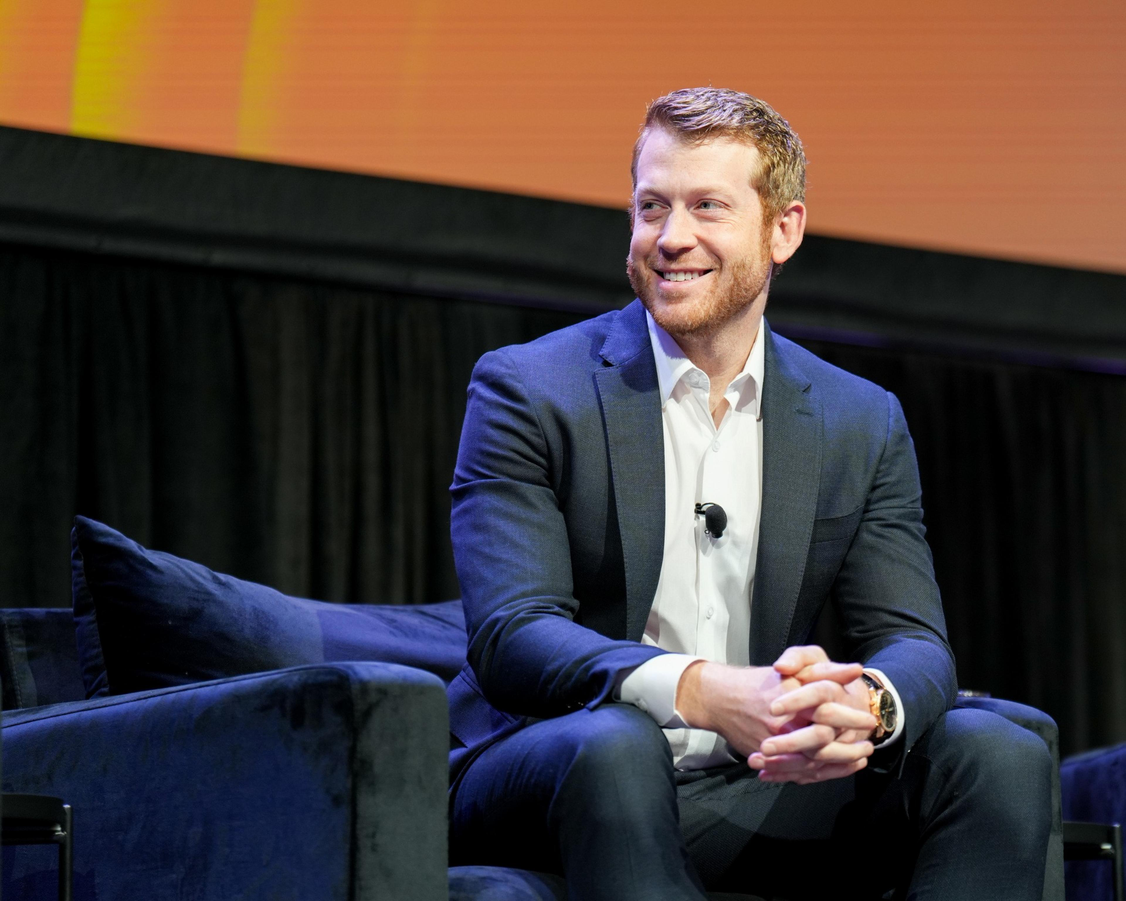 A man in a dark suit sits a dark chair against an orange-and-black backdrop.