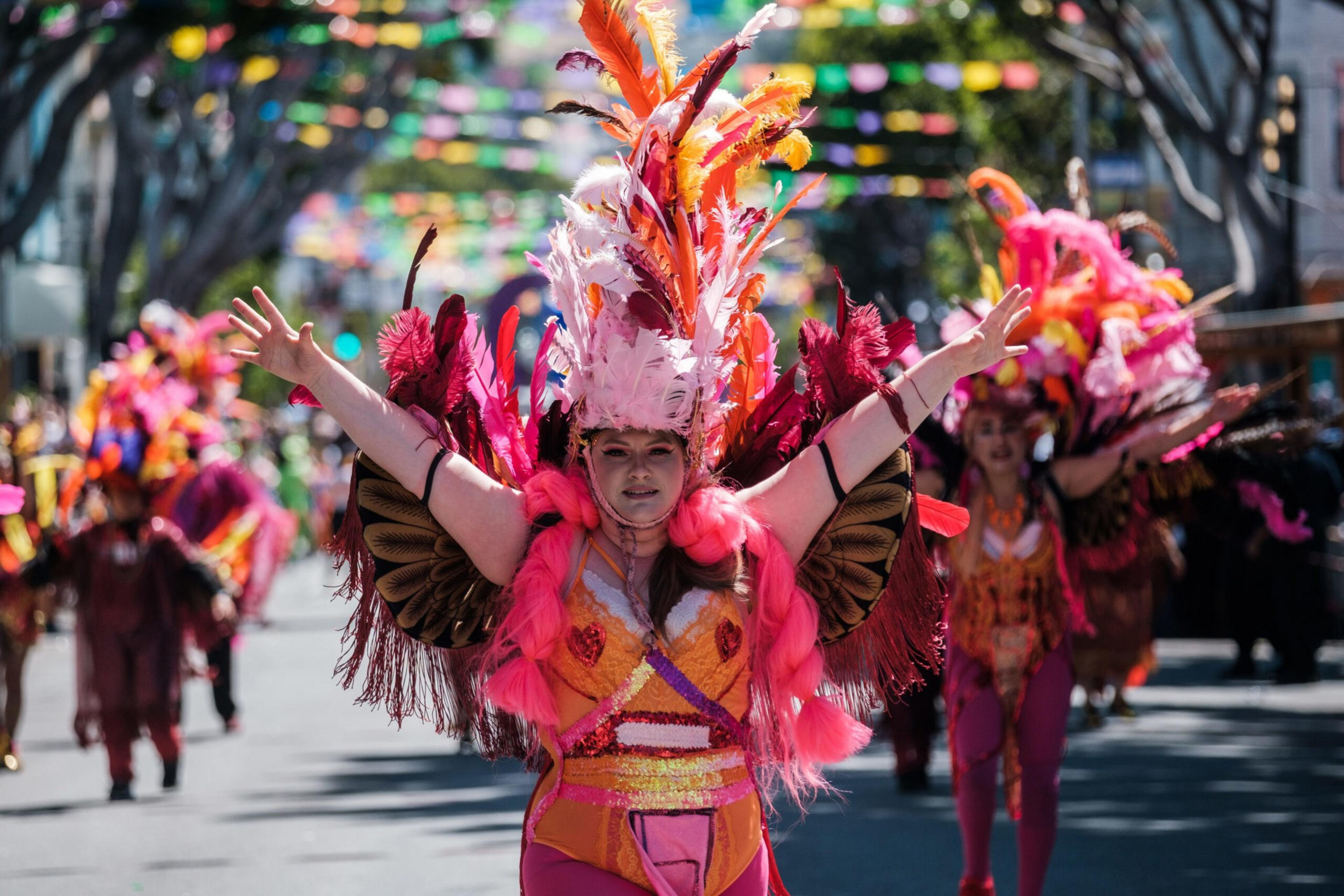 a woman in an elaborate red costume and feathers sticking out of her cap with her arms outstretched dances in a street with people in similar outfits