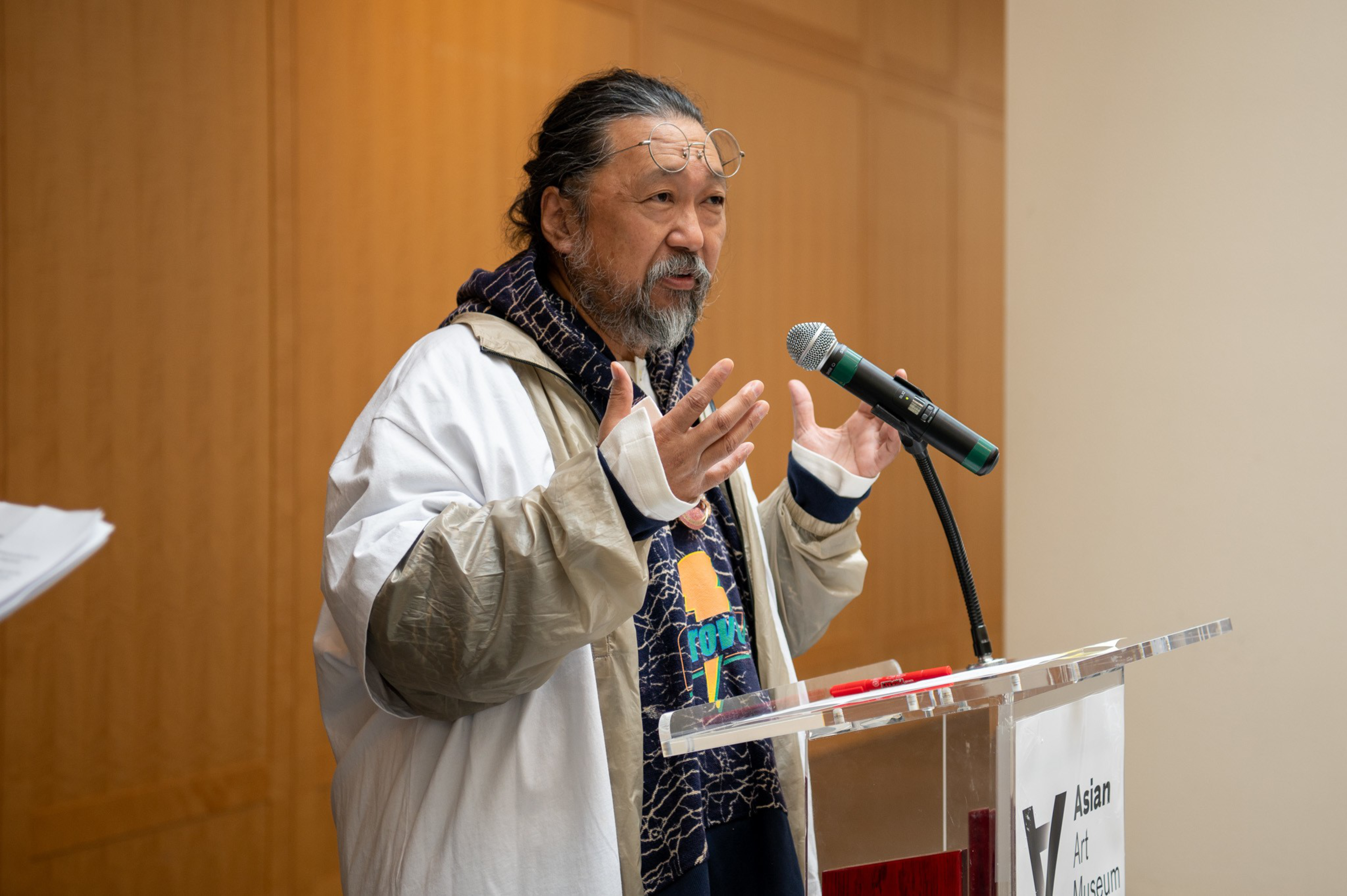 A man with a beard and glasses speaks at a podium with a microphone, wearing a patterned hoodie under a light jacket inside a room.