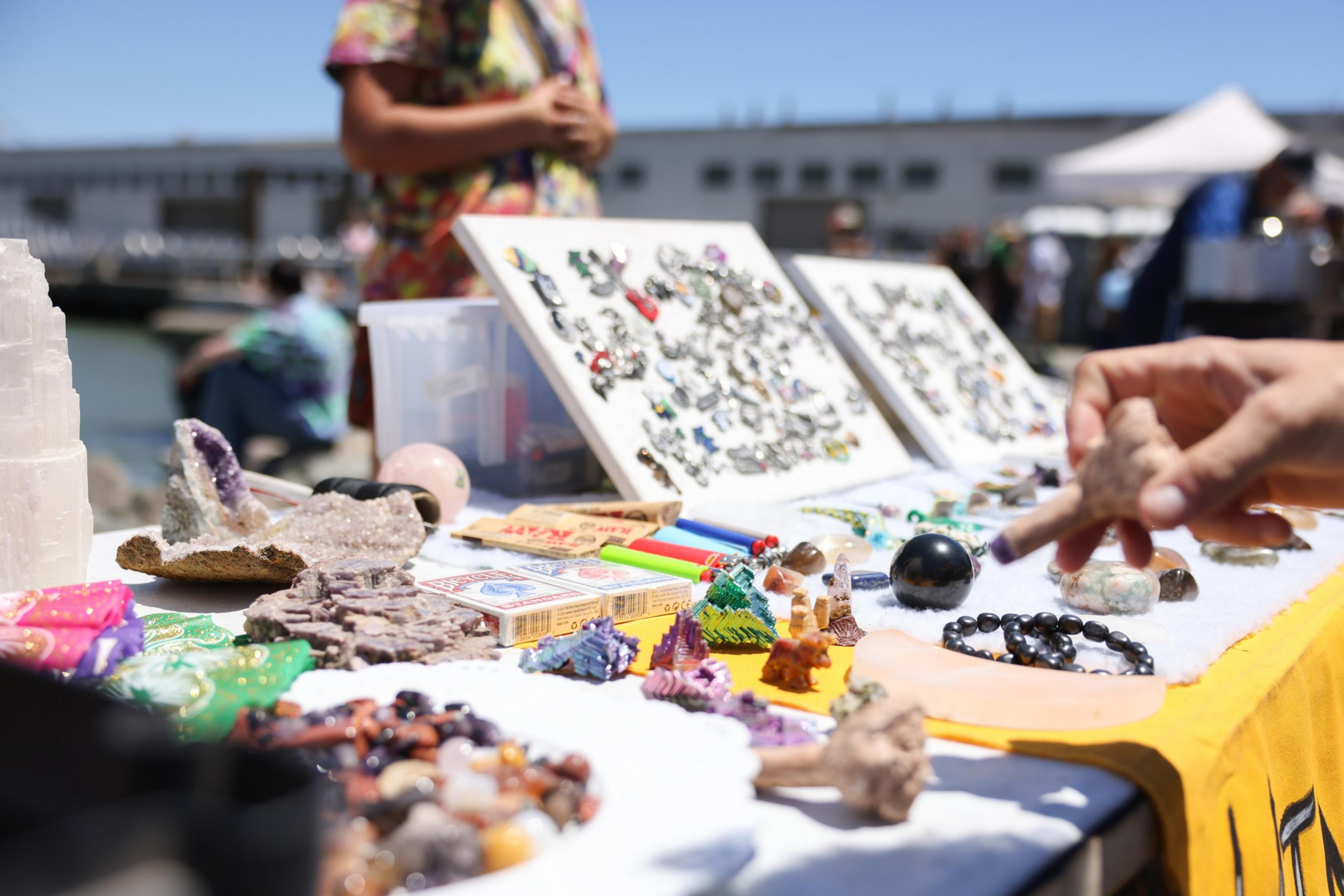 A table displays various colorful crystals, stones, jewelry, and trinkets under sunlight. A person’s hand touches an item while others browse the outdoor market.