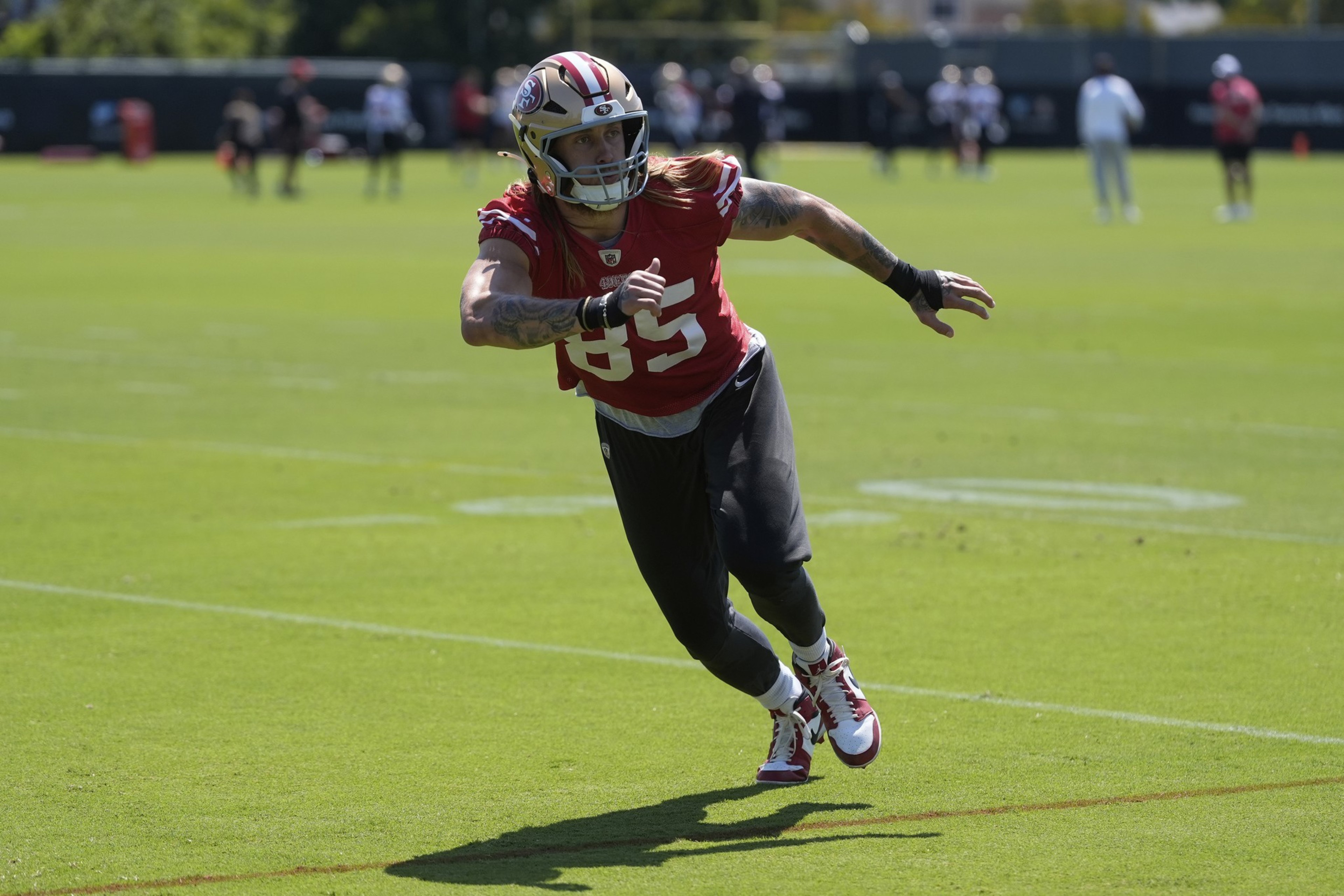 A football player in a red jersey and black pants is running on a green field during practice. He's wearing a helmet and has tattoos on his arms.