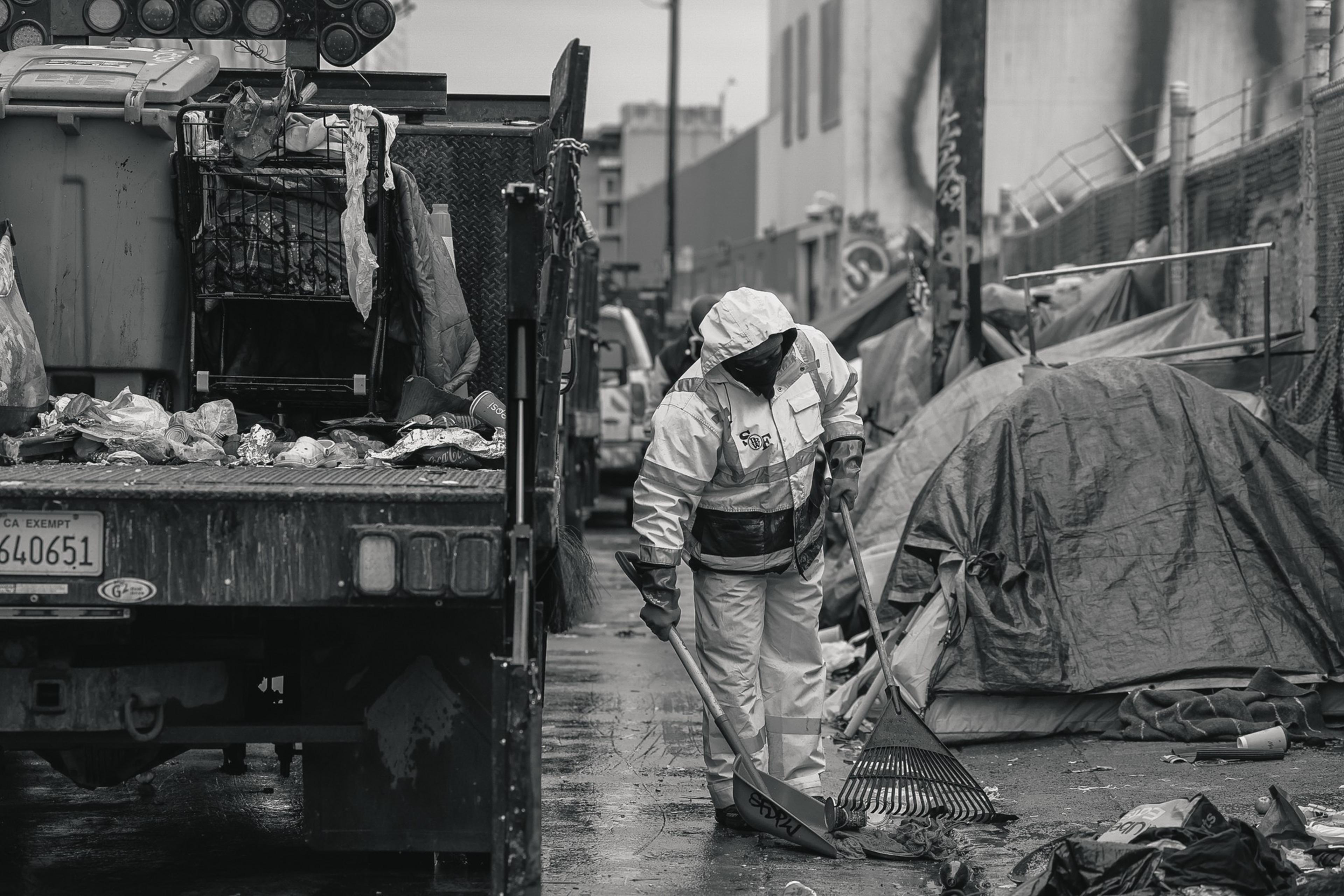 A worker in a bright yellow rain suit uses a shovel and rake to clean a trash-littered street beside a sanitation truck and a makeshift tent encampment.