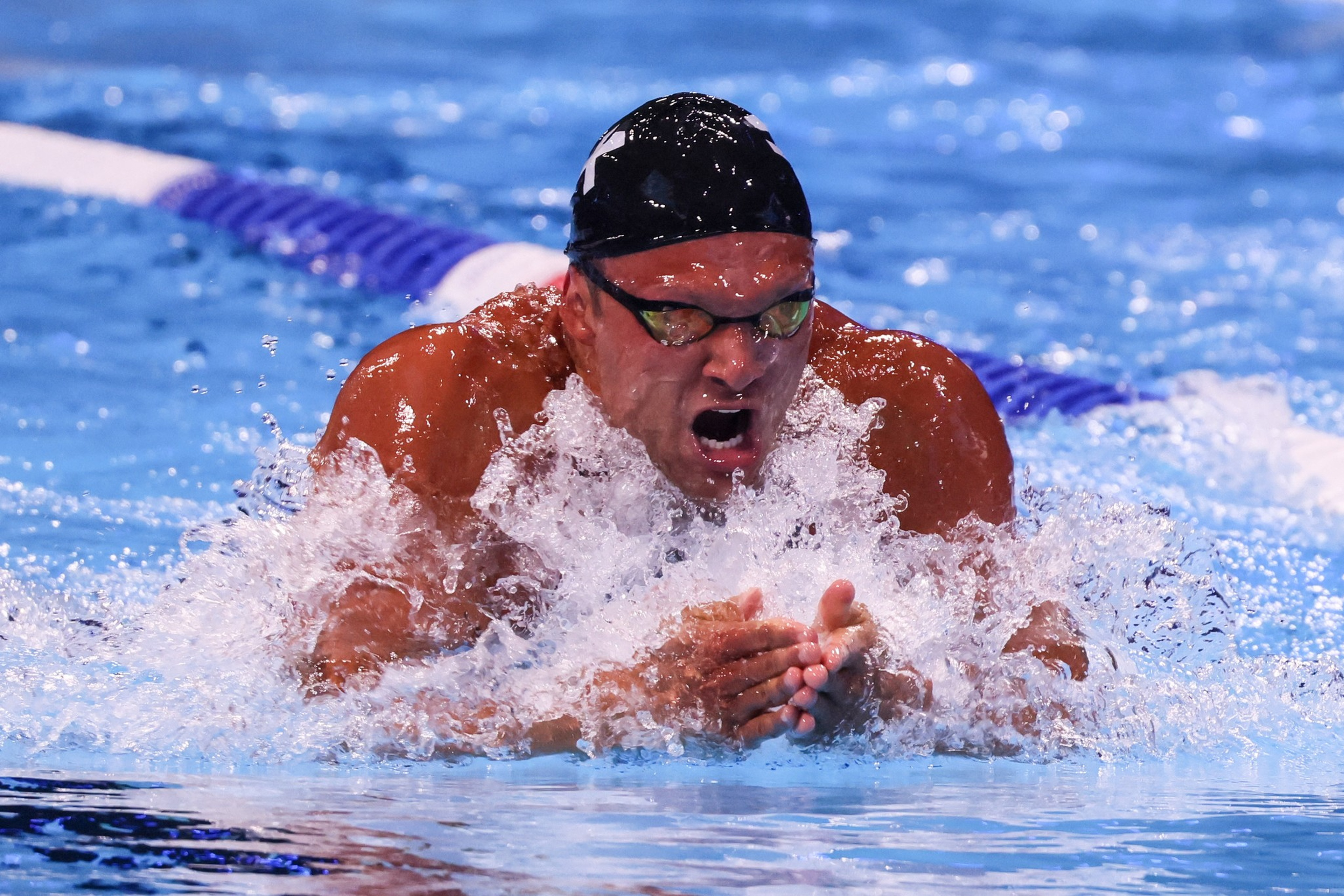 A swimmer wearing a black cap and goggles is intensely swimming breaststroke, creating splashes in a pool lane marked with a purple divider.