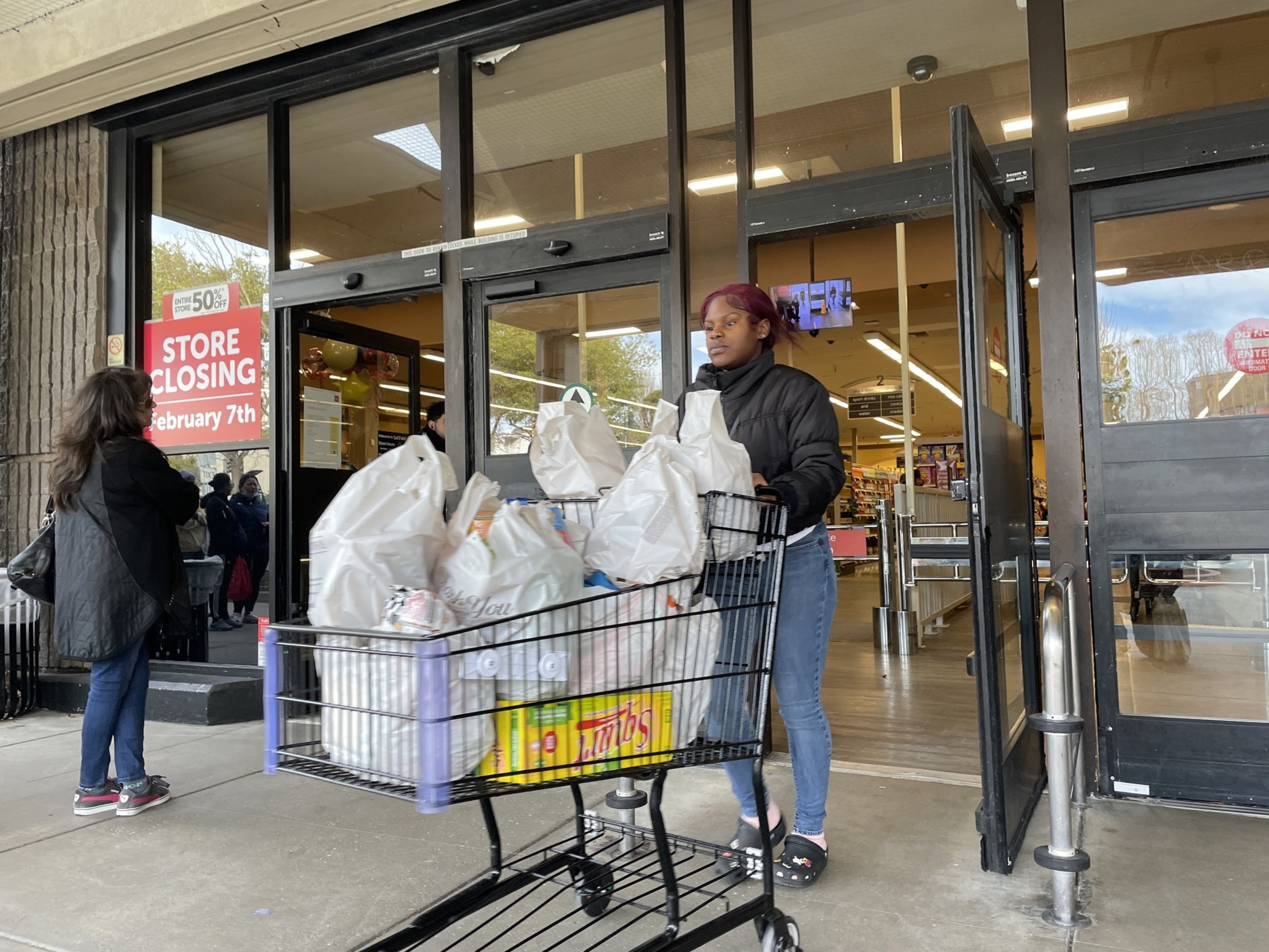 A woman pushes a shopping cart filled with bags from a store. A sign on the window announces &quot;Store Closing February 7th.&quot; Another person stands nearby.