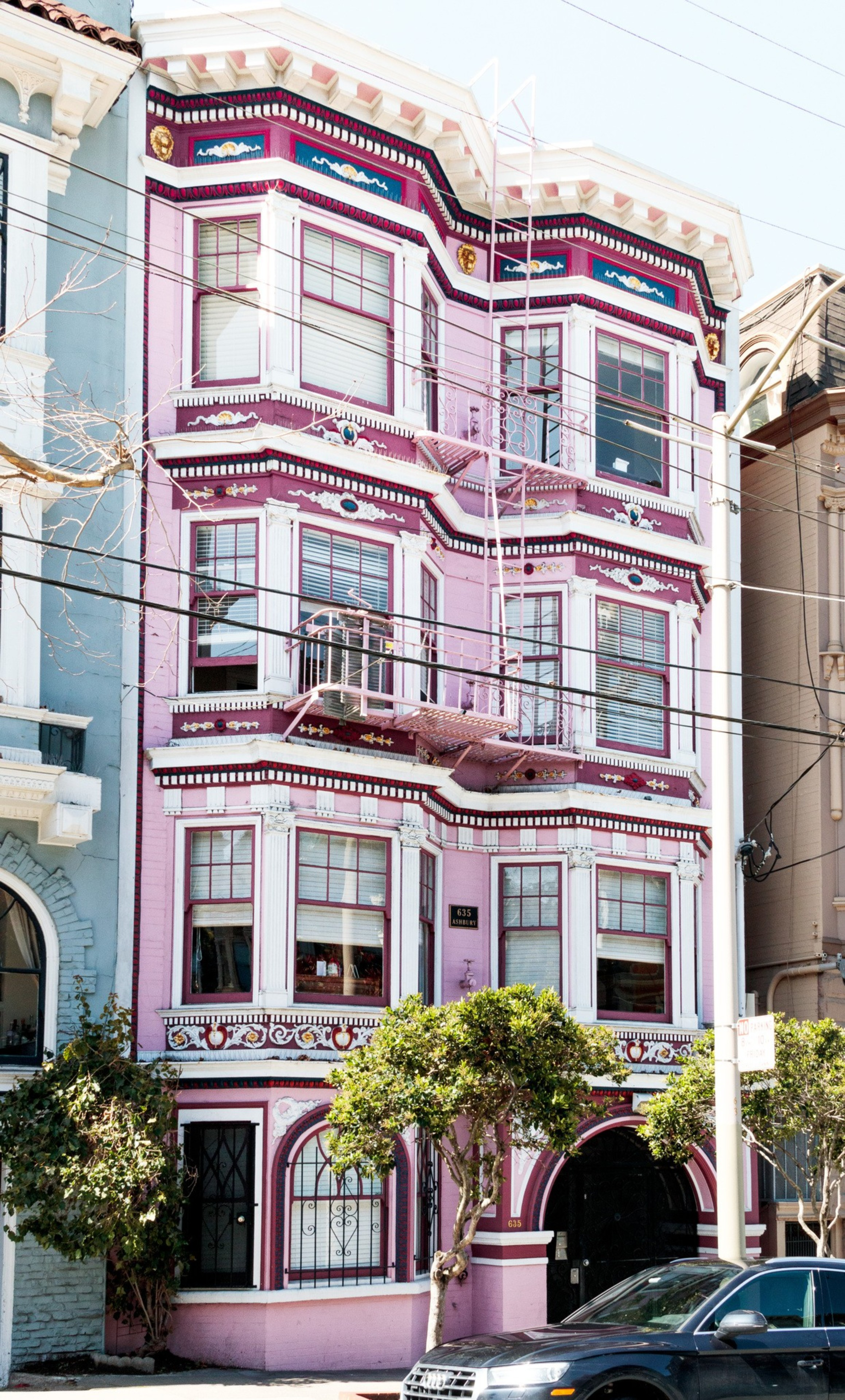 A three-story pink Victorian building with ornate white and purple trim, bay windows, a fire escape, trees in front, and a parked black car.