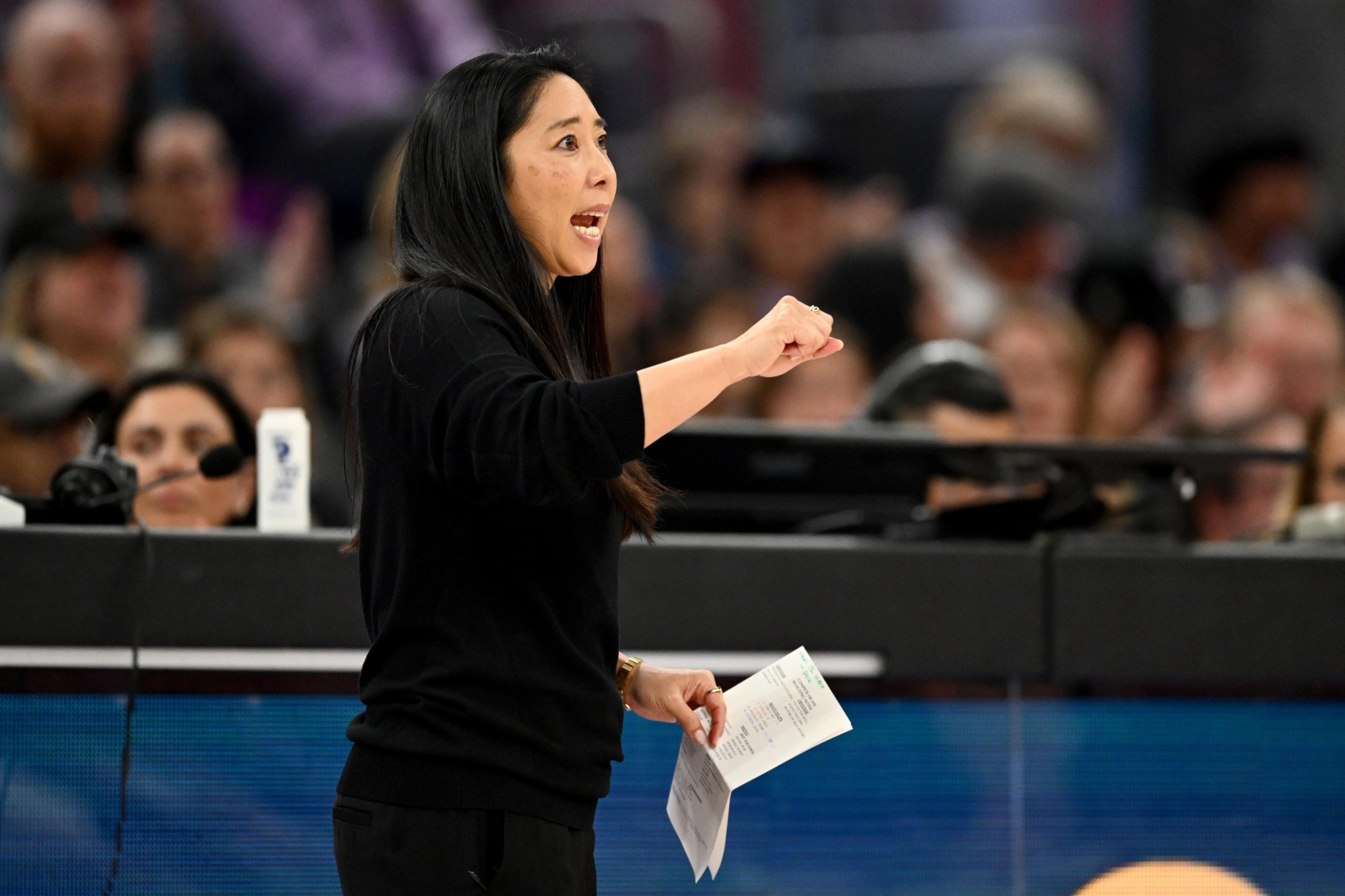 A woman with long dark hair, in a black sweater, is speaking energetically while holding papers. A crowd is in the blurred background.
