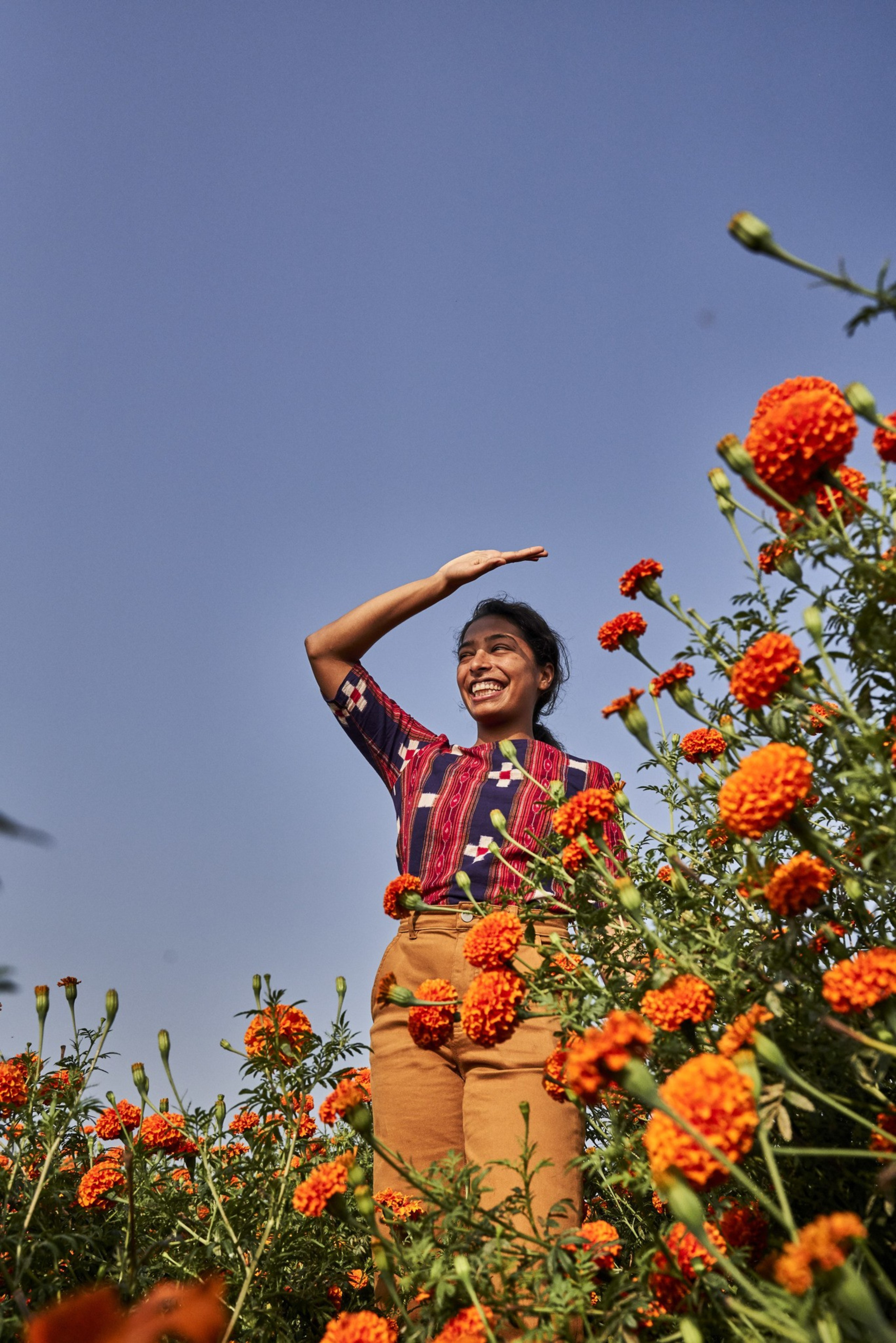 A smiling woman stands among vibrant orange marigold flowers, shading her eyes with one hand against a clear blue sky.