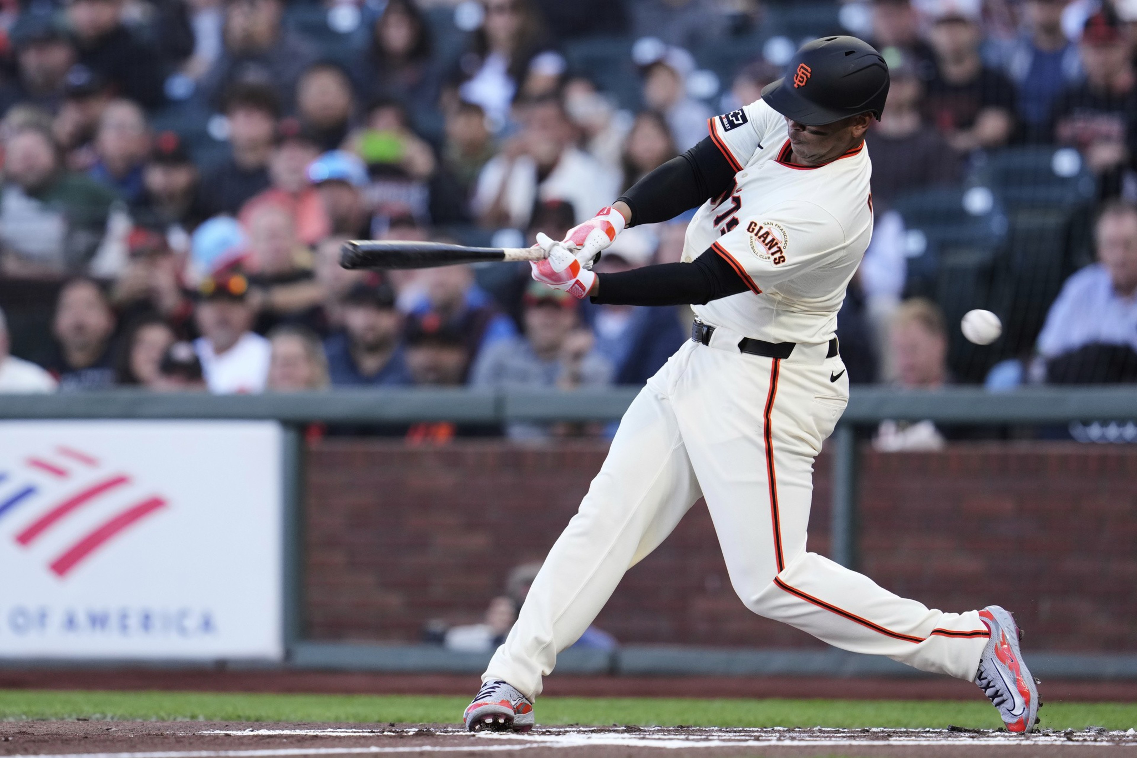 A baseball player in a white uniform is swinging a bat, likely hitting the ball. He's in a stadium with a blurred audience in the background.