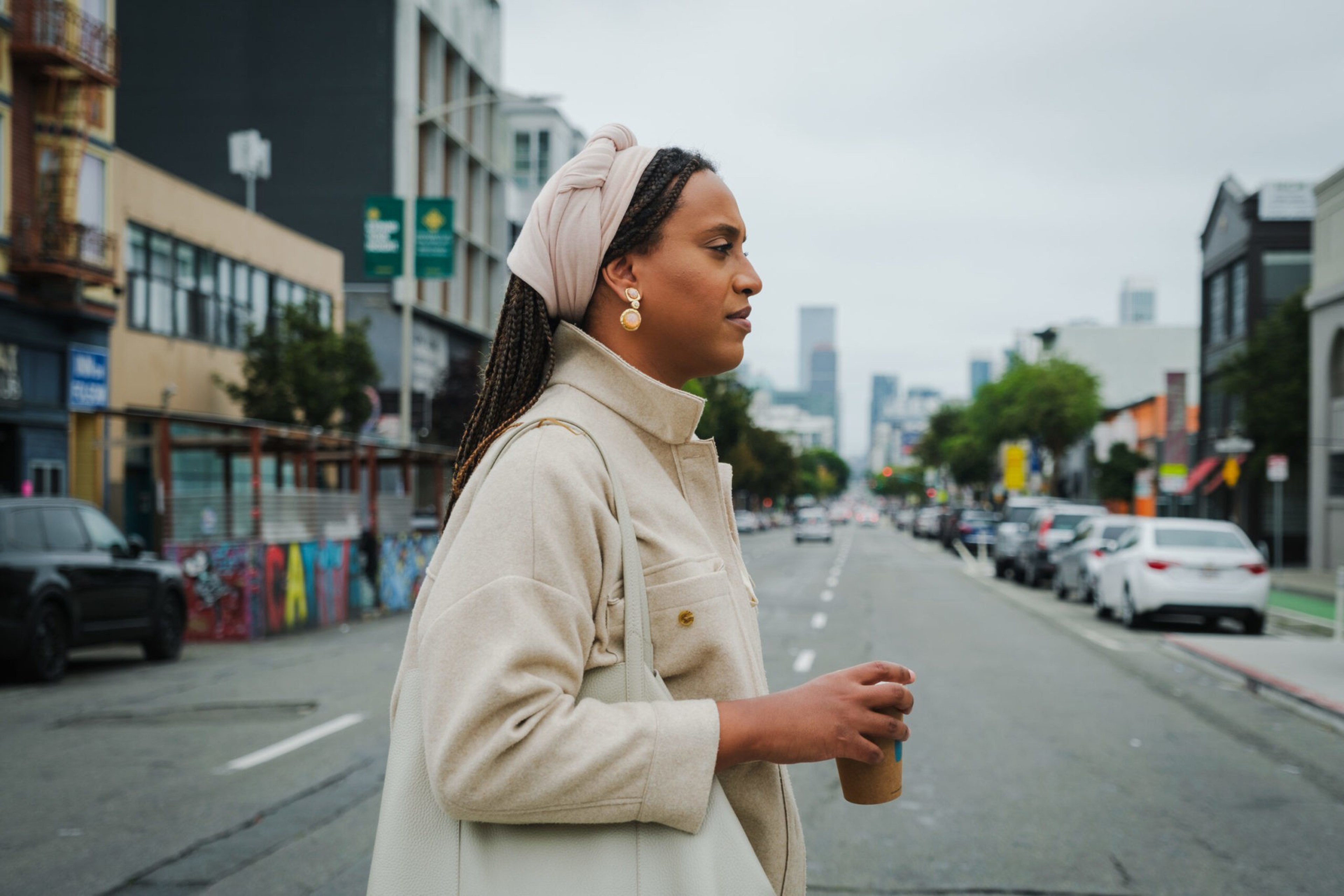 Honey Mahogany crosses the street wearing a white coat and carrying a cup of coffee.
