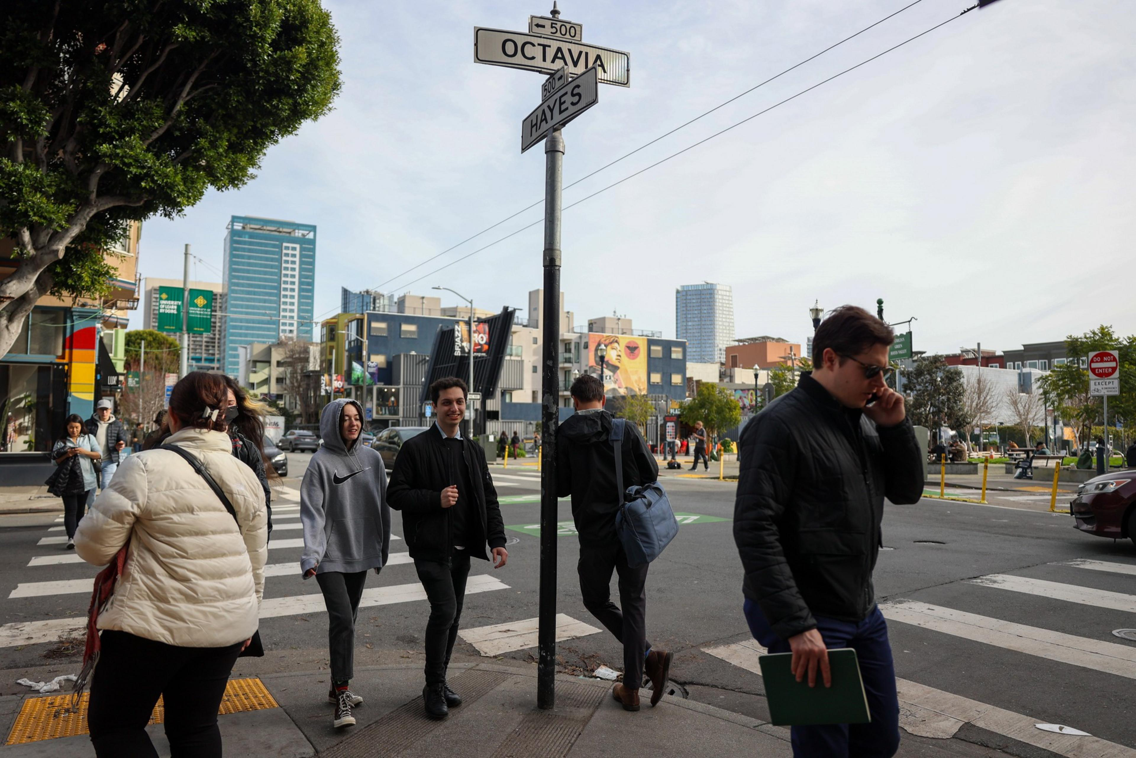 A busy street intersection shows people walking in various directions. Some are crossing, while one person talks on the phone. Buildings and street signs are visible.