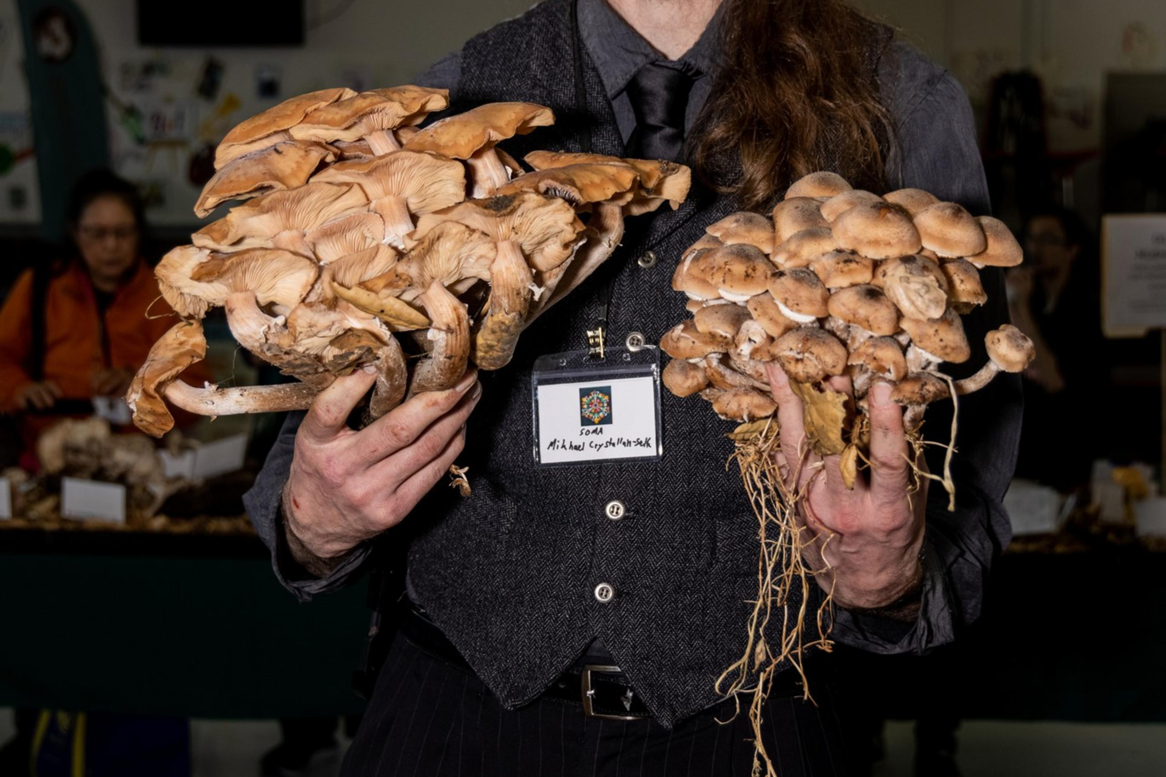 A man with long hair and glasses holds two large clusters of mushrooms, wearing a dark vest and tie. The room in the background has people and tables.