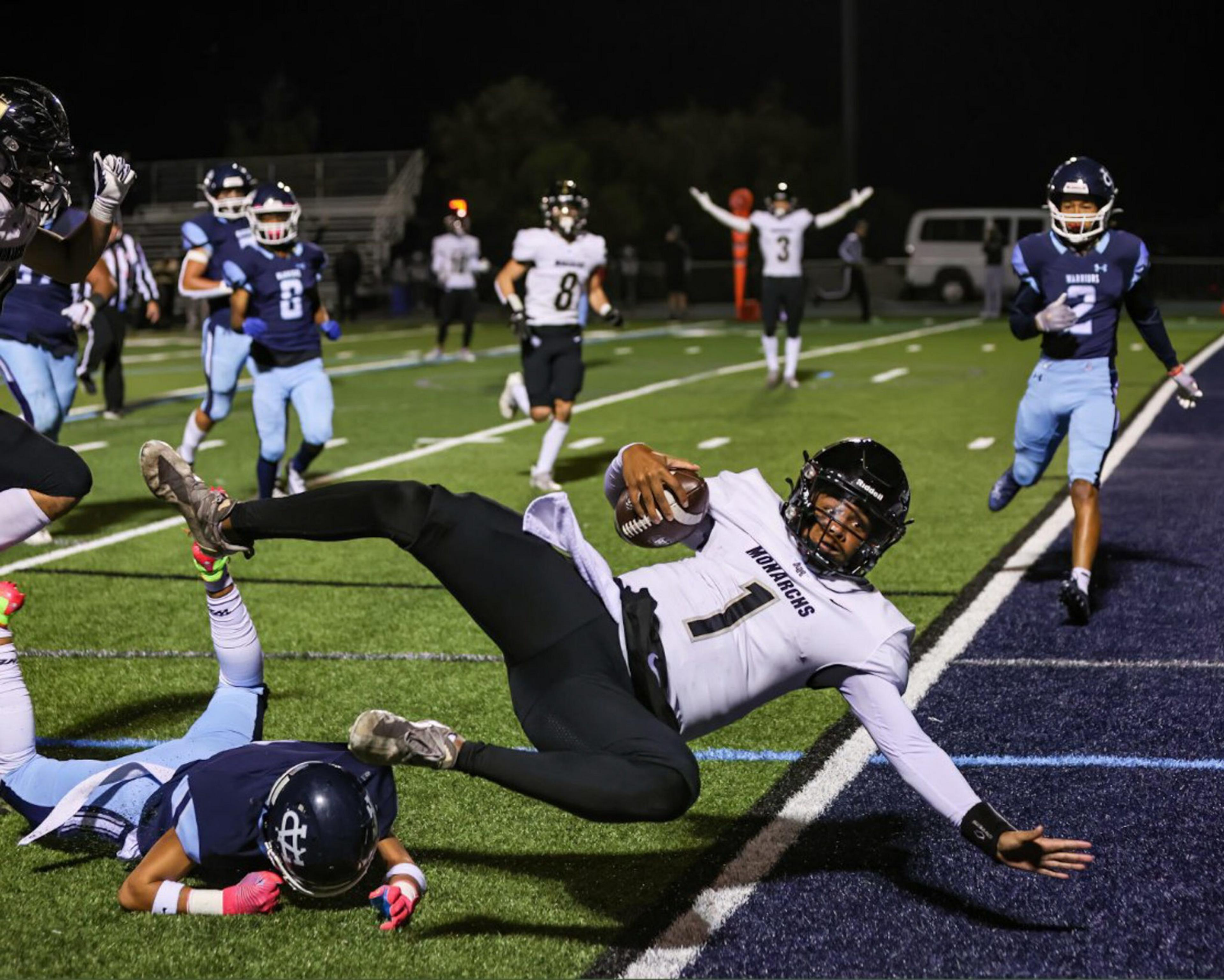 A football player in a white jersey dives toward the end zone while a blue-jerseyed defender falls to the ground behind him.