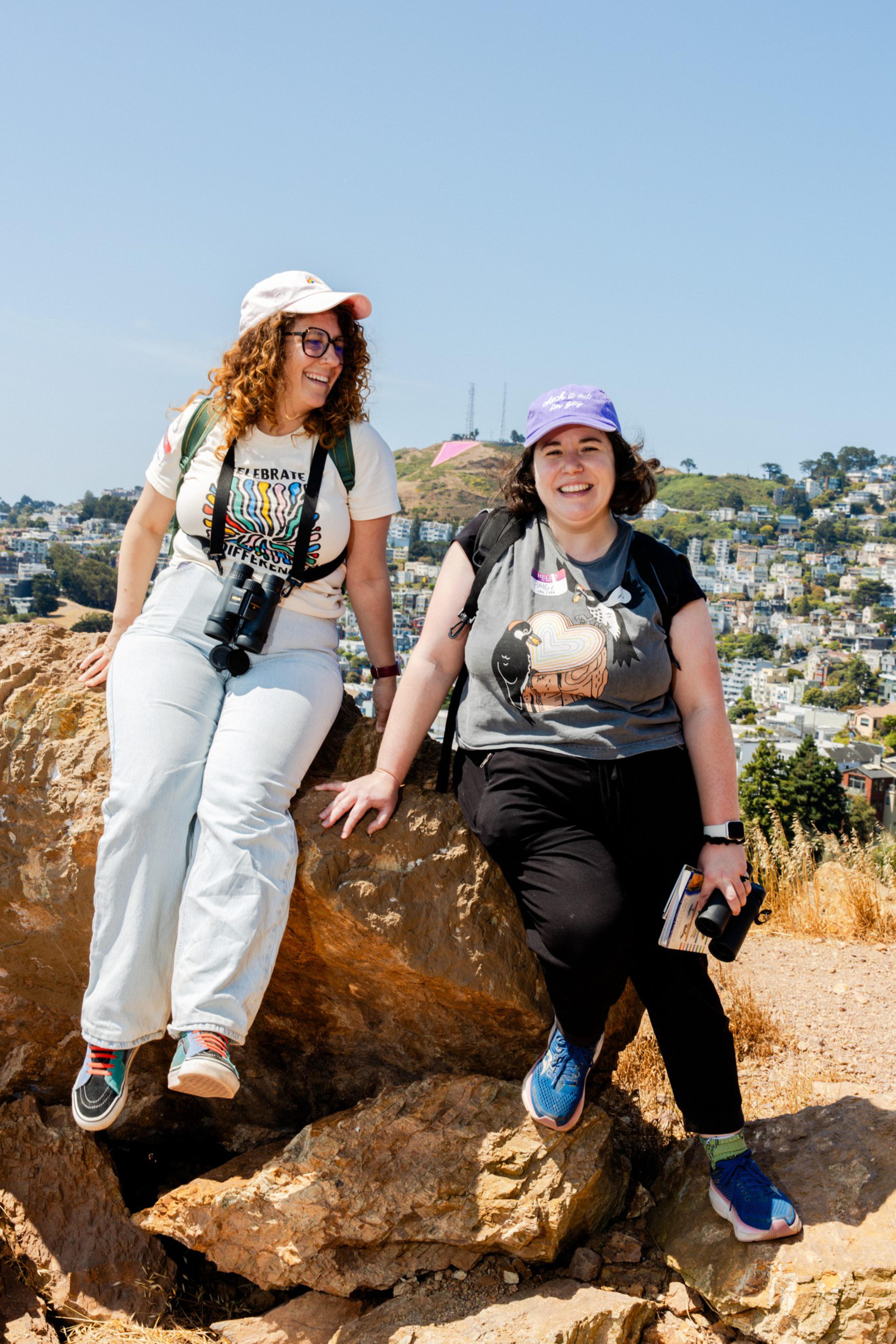 Two people are sitting on rocks, smiling. They wear casual outdoor clothes, hats, and hold binoculars. A hilly urban landscape is visible in the background.