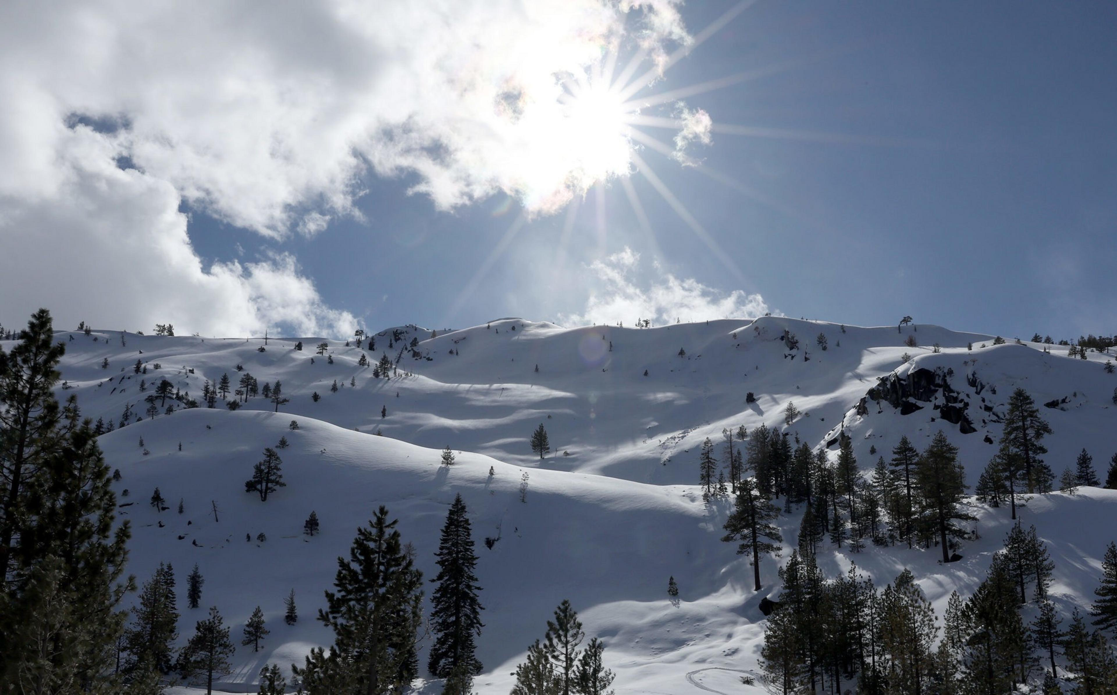 Snow-covered hills with scattered pine trees under a bright sun partially hidden by clouds in a clear blue sky.