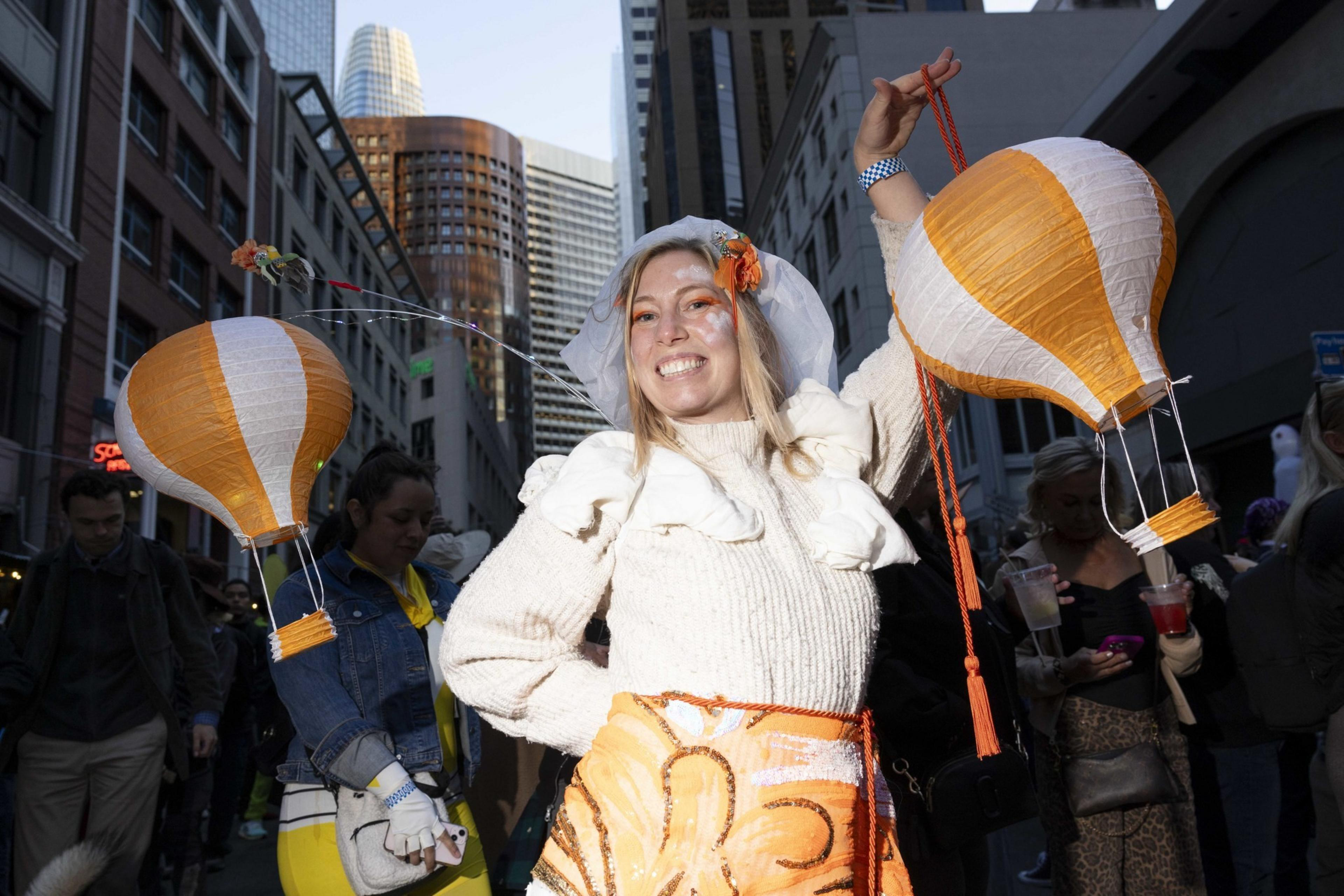 A smiling person in an urban setting holds two orange and white balloon-shaped lanterns. They wear a white top with decorative elements and are surrounded by people.