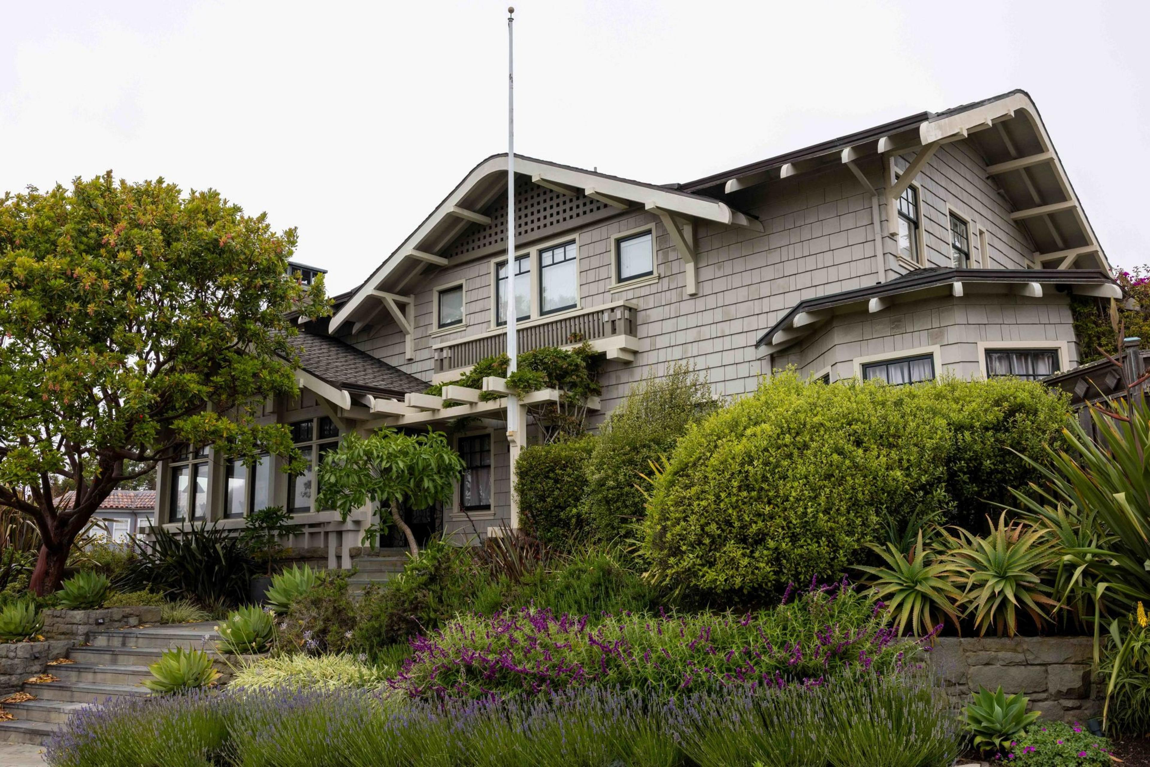 A charming gray house with a sloped roof is surrounded by lush green bushes, colorful flowers, and a well-maintained garden path leading to the entrance.