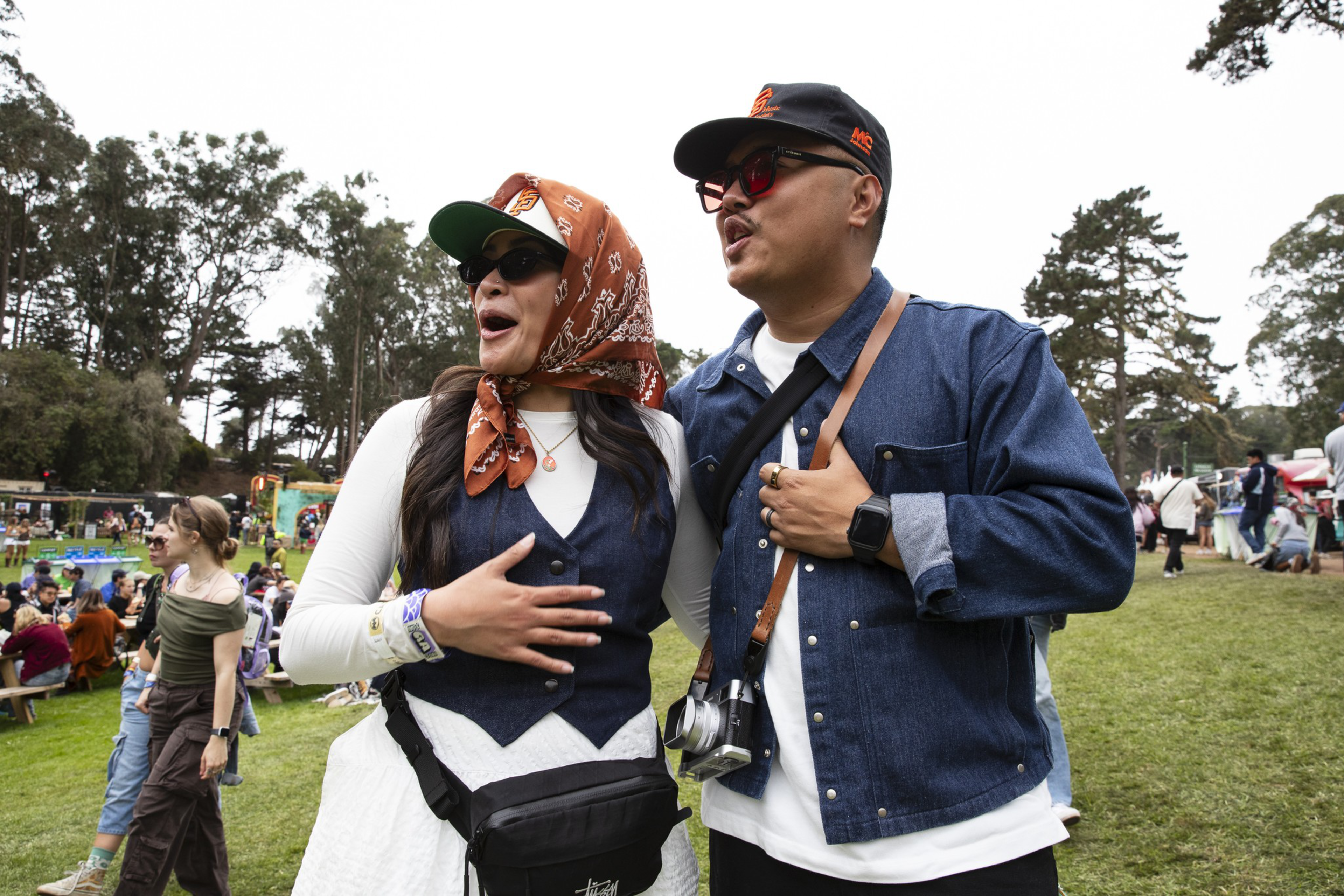 A man and woman wearing sunglasses and casual outdoor clothes stand close, looking to the side with surprised or excited expressions at a park event.