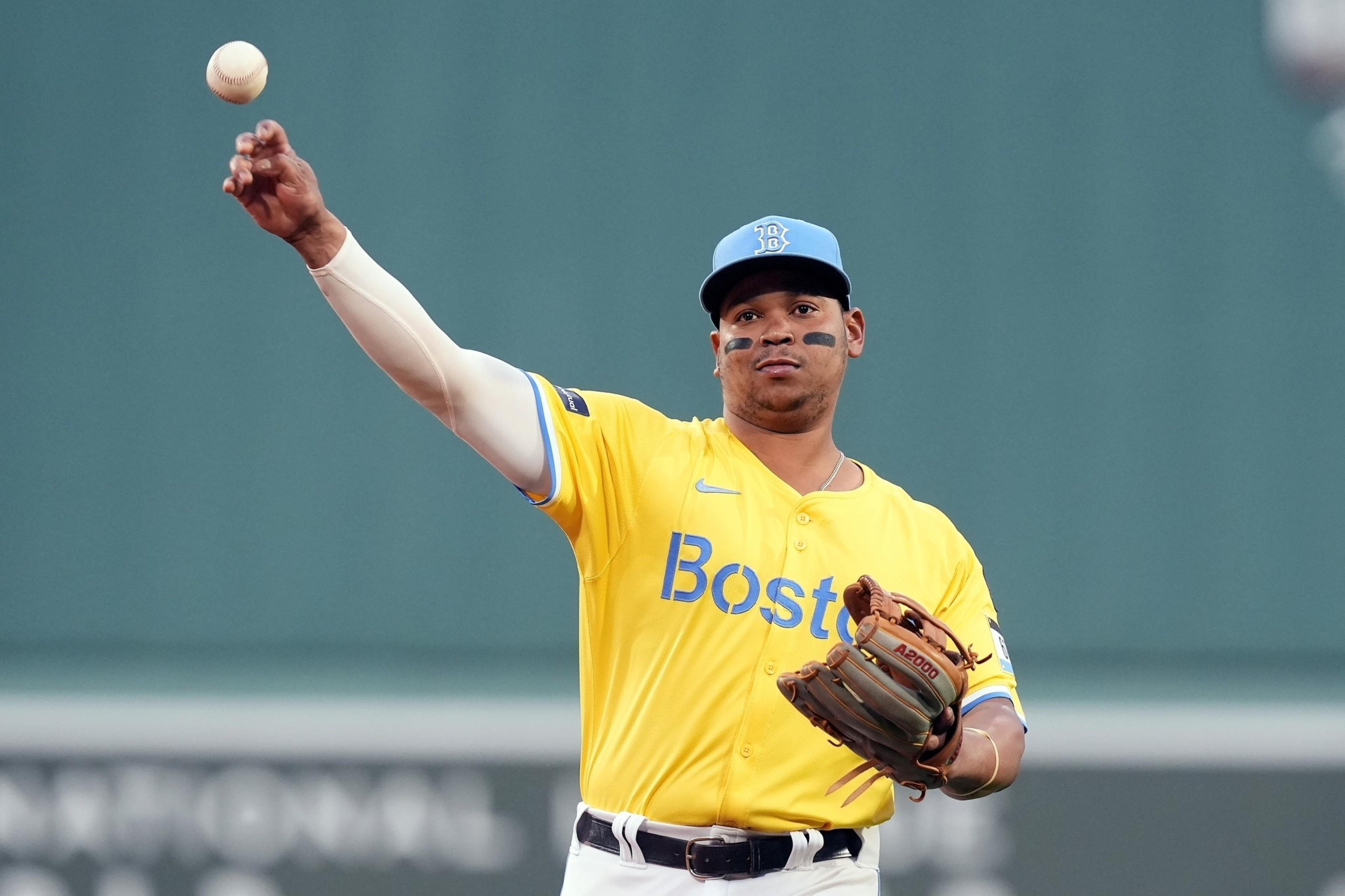 A baseball player in a yellow &quot;Boston&quot; jersey and blue cap throws a ball. He wears a glove on his left hand, and has black eye paint under his eyes.