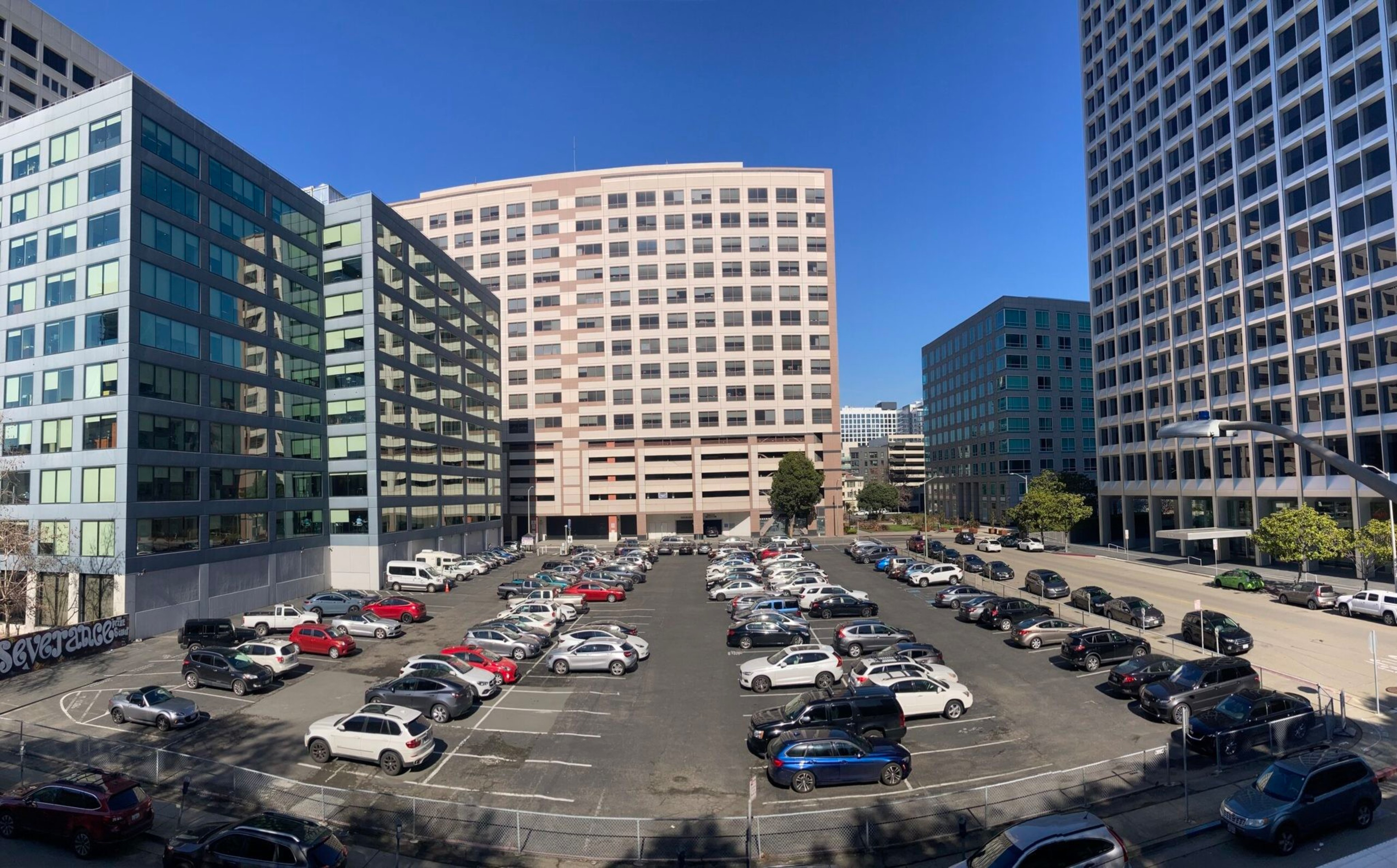 A nearly full outdoor parking lot is surrounded by tall office buildings under a clear blue sky.