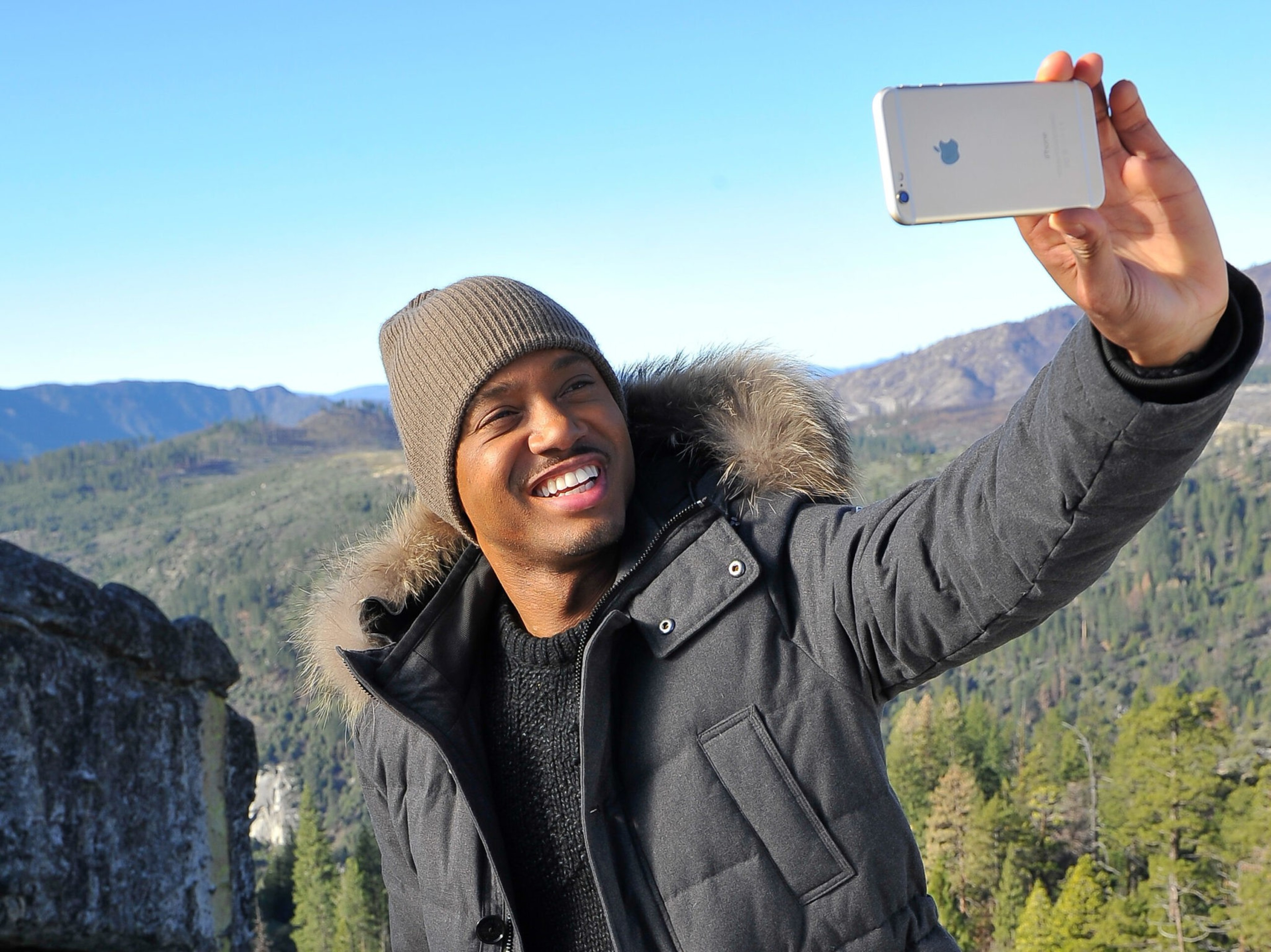 A smiling man in a winter coat and knit hat takes a selfie with a smartphone against a backdrop of mountains and evergreen trees.