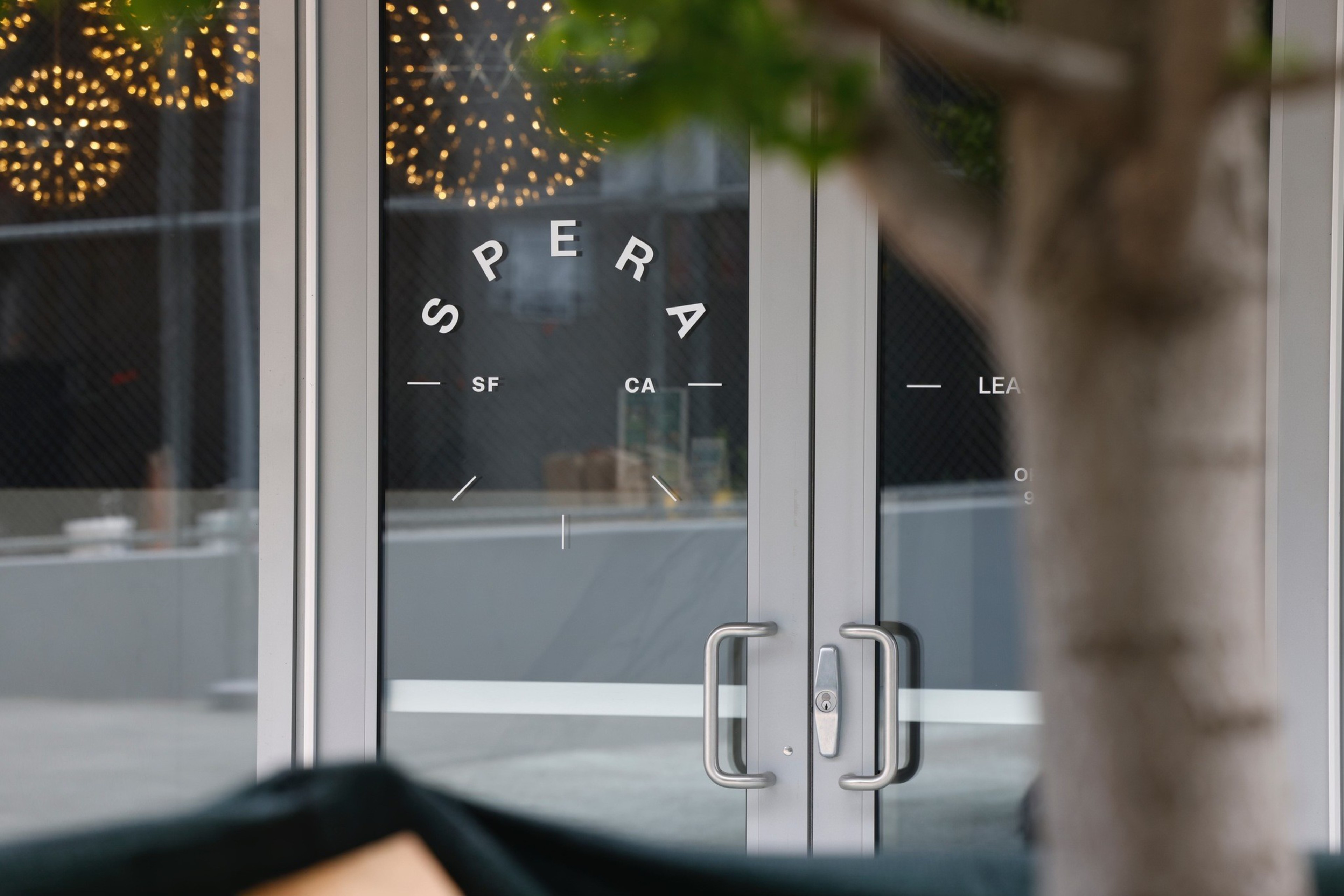 A glass door features modern decor with the word &quot;SPERA&quot; and the letters &quot;SF&quot; and &quot;CA&quot;. Decorative hanging lights are seen through the door; a tree is in the foreground.