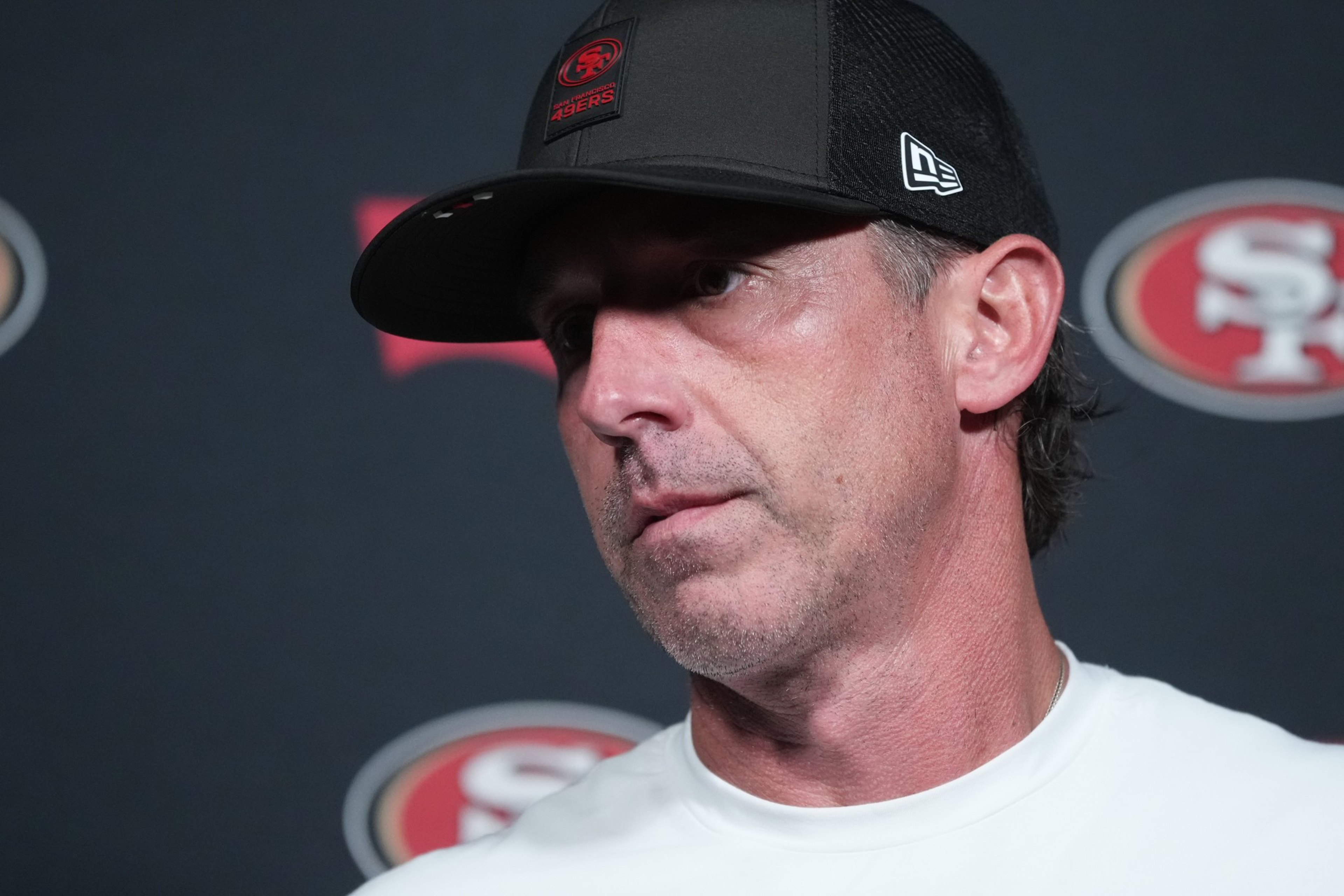 A man wearing a black San Francisco 49ers cap and white shirt stands against a backdrop featuring the team’s logo.