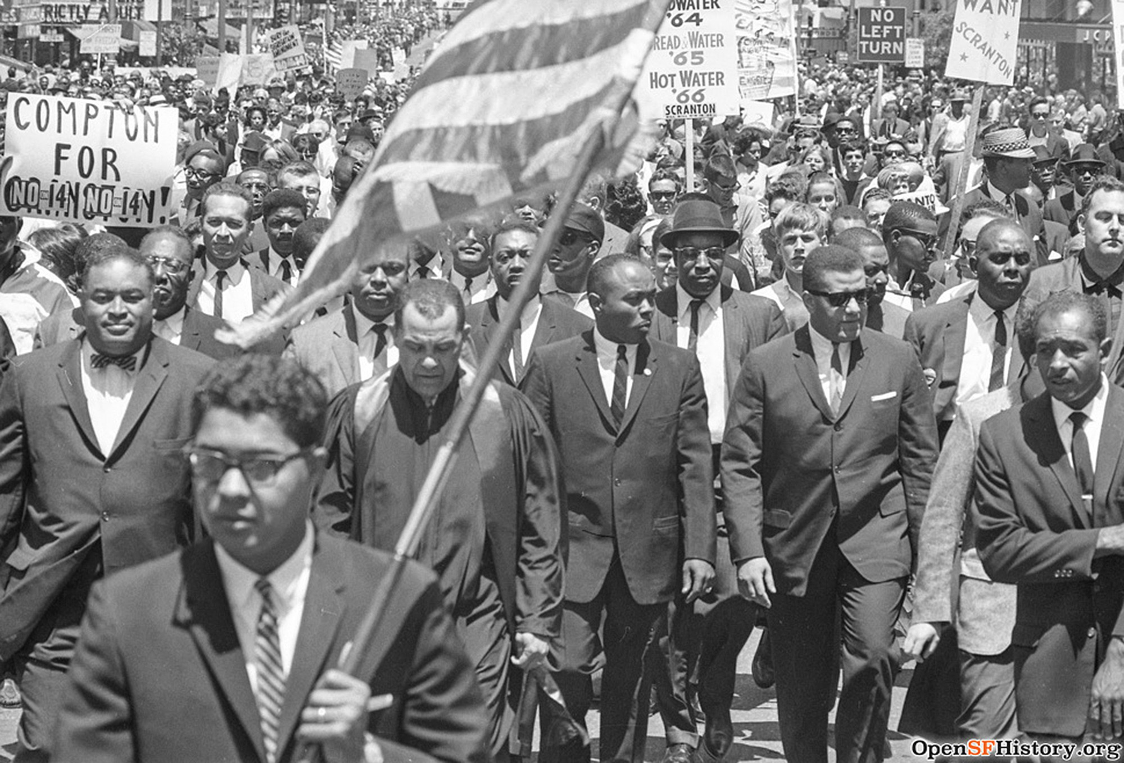 A large, diverse crowd of men in suits march in a civil rights protest, holding signs and an American flag.