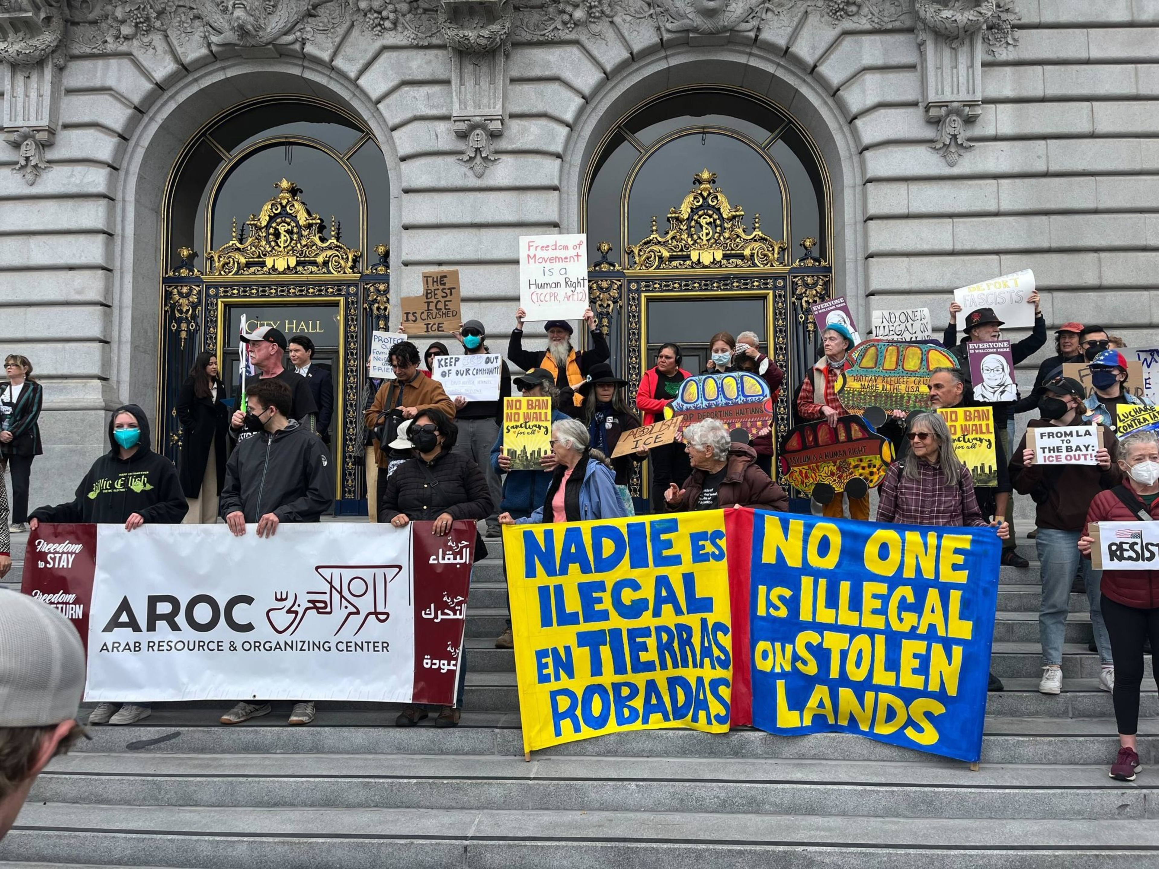 A group of people stand on steps holding signs with messages about freedom and immigration, in front of ornate City Hall doors.