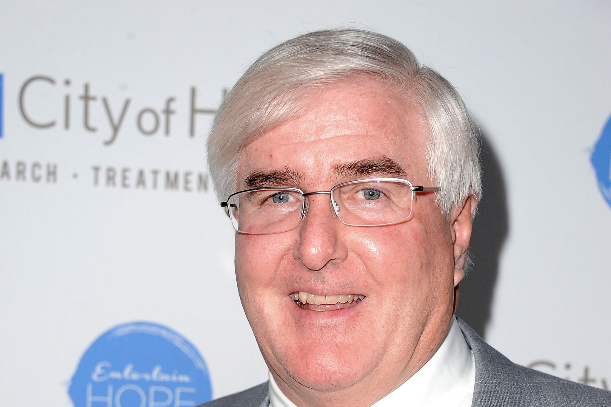 A man with short, white hair, glasses, and a grey suit with a light blue tie smiles at the camera. The backdrop has the text "City of Hope" and a blue logo.