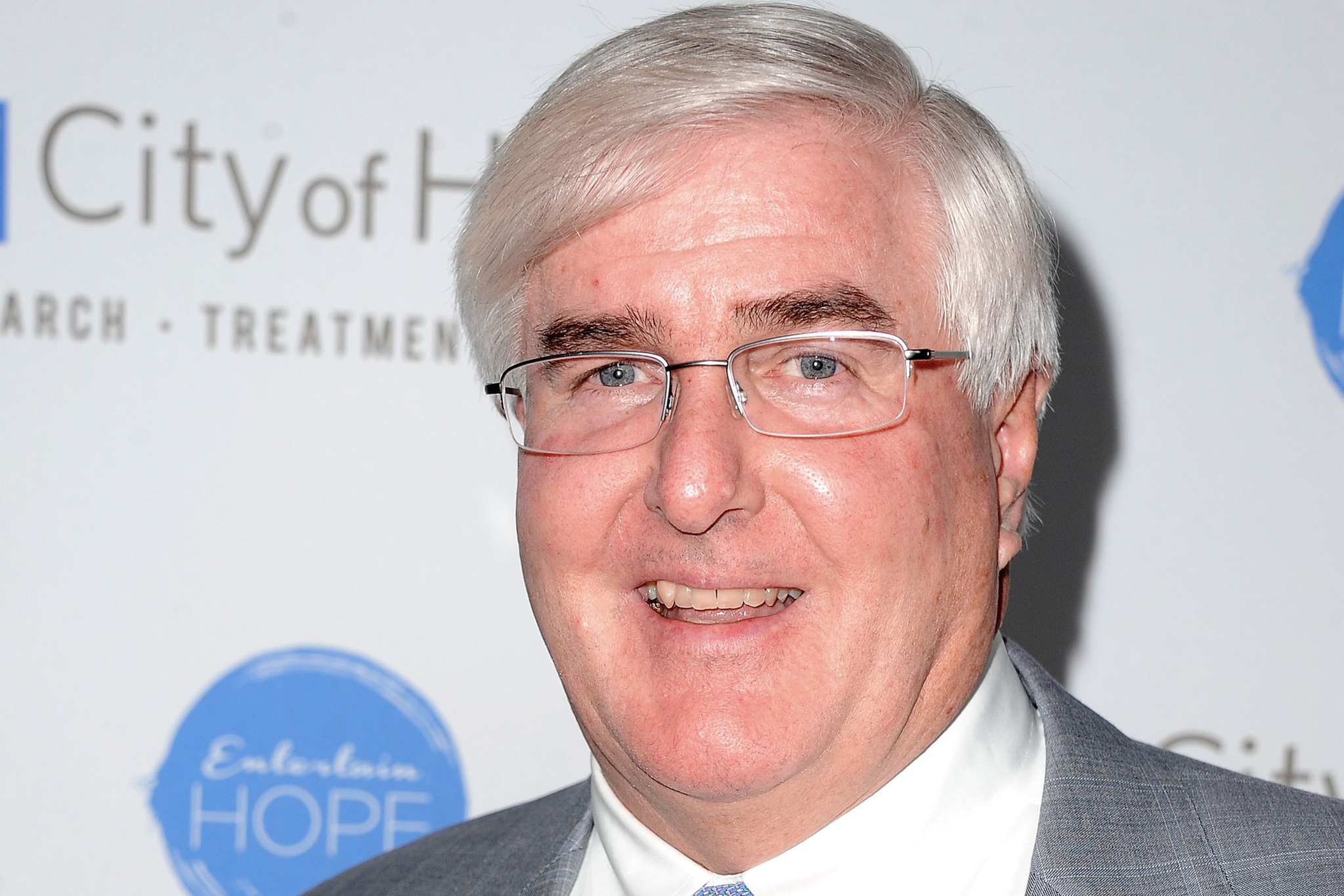 A man with short, white hair, glasses, and a grey suit with a light blue tie smiles at the camera. The backdrop has the text "City of Hope" and a blue logo.
