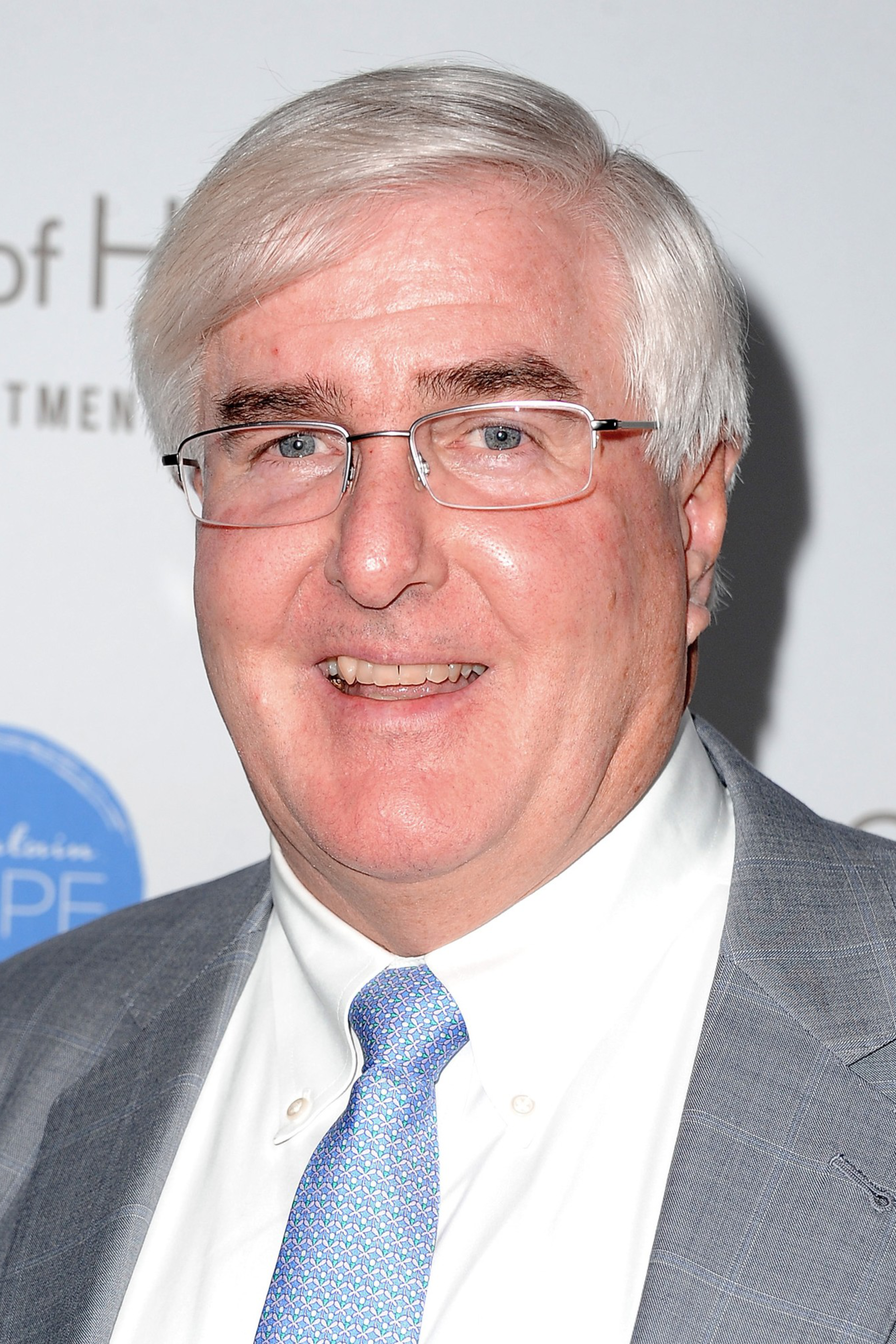 A man with short, white hair, glasses, and a grey suit with a light blue tie smiles at the camera. The backdrop has the text "City of Hope" and a blue logo.