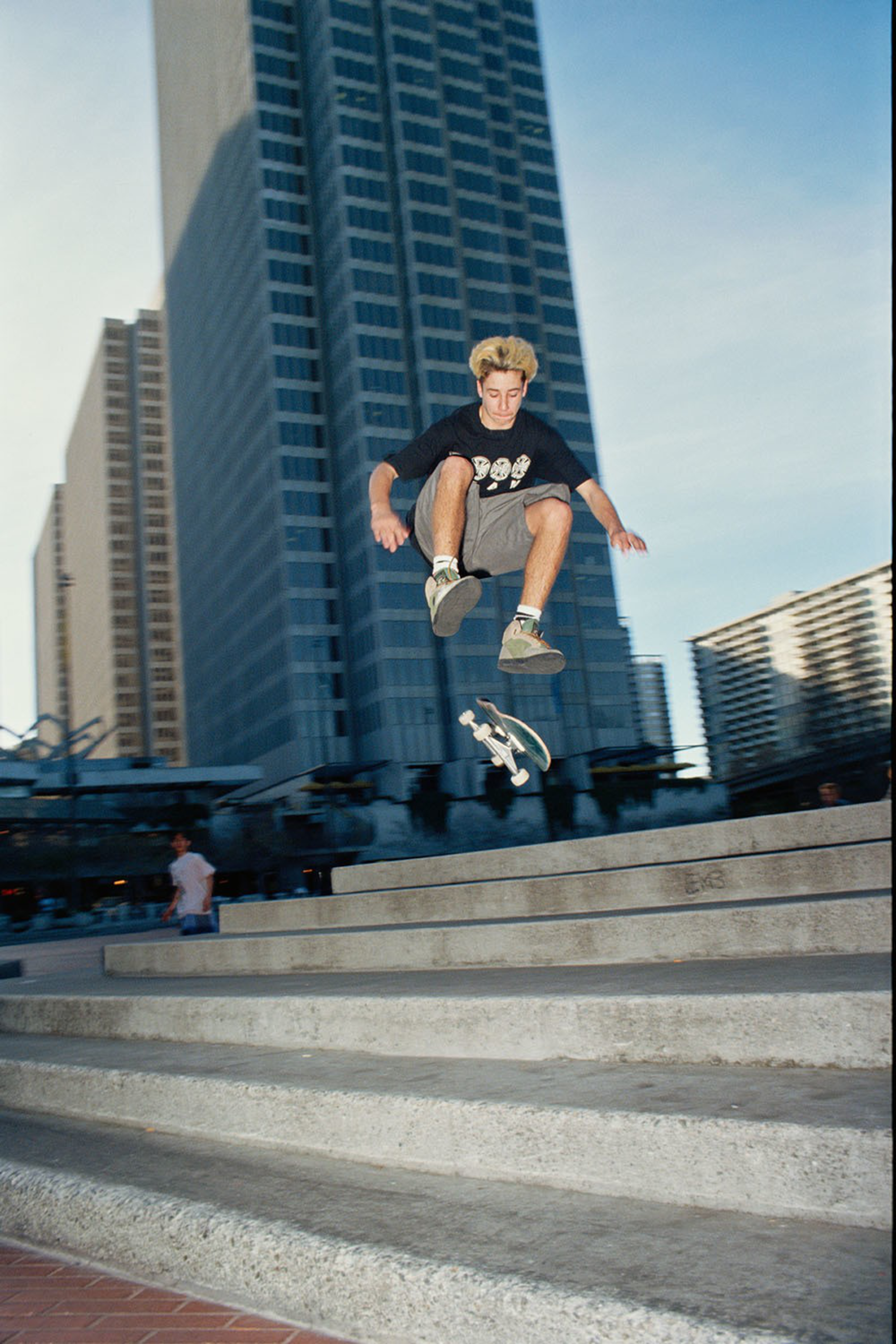 A young skateboarder performs a mid-air trick over a set of concrete stairs with tall buildings in the background.