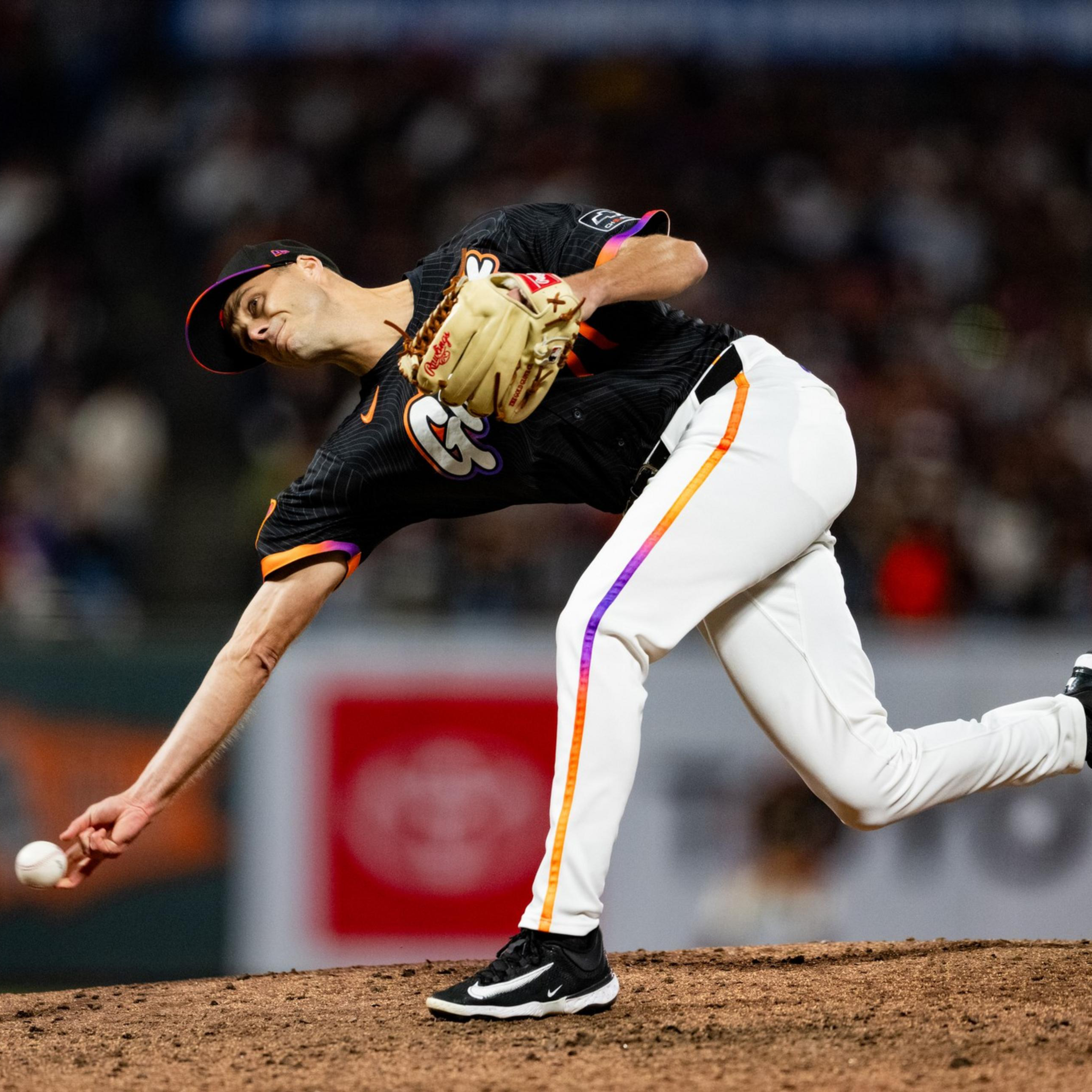 A baseball pitcher in a black jersey and white pants is releasing a pitch from the mound during a nighttime game.