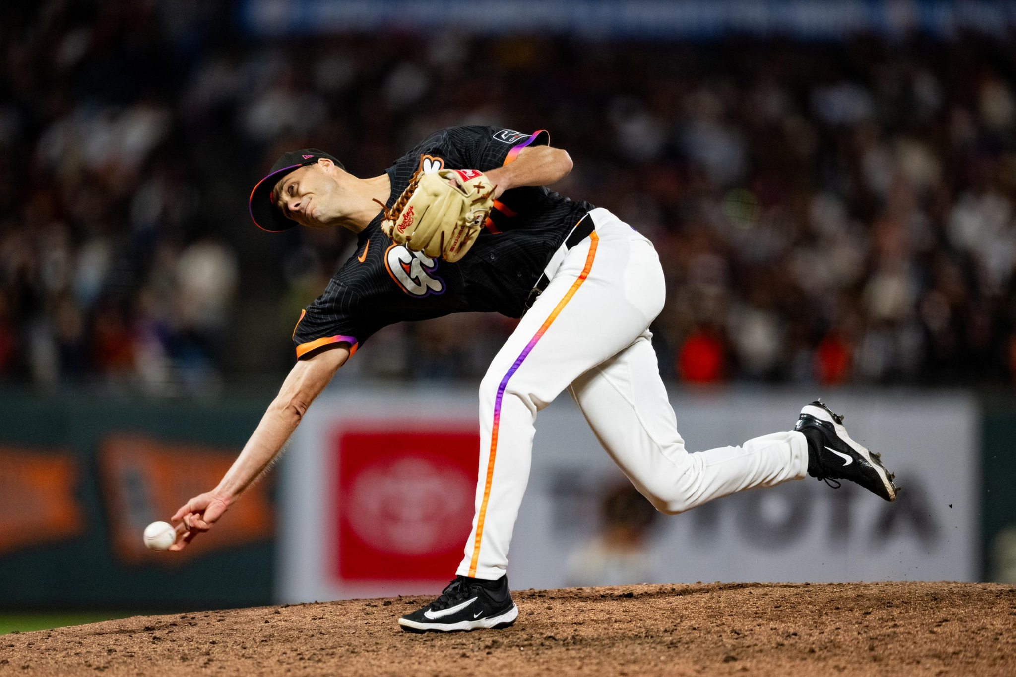 A baseball pitcher in a black jersey and white pants is releasing a pitch from the mound during a nighttime game.