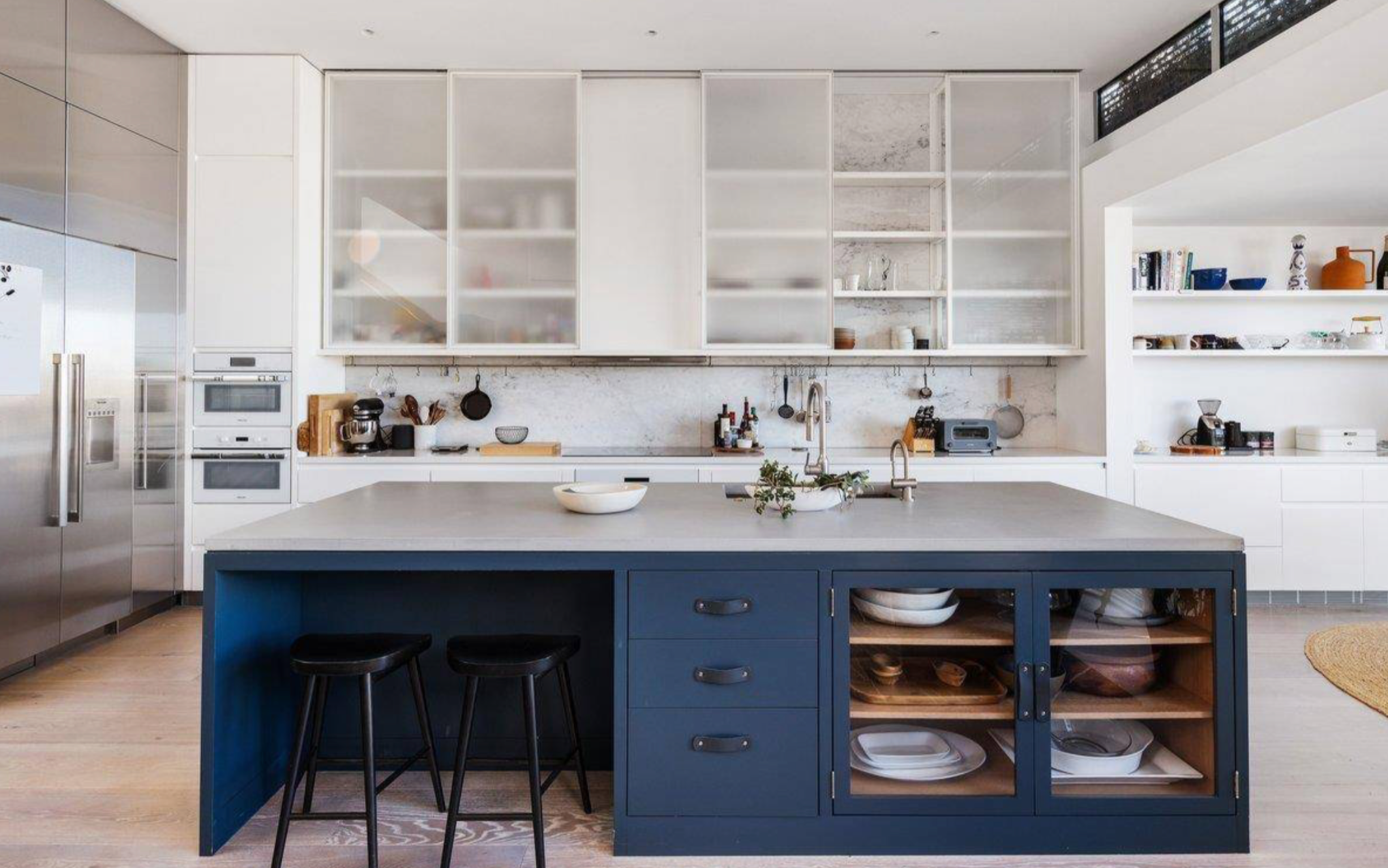 A modern kitchen with a large blue island featuring stools and storage, white frosted cabinets, stainless steel appliances, and open shelves with dishes and decor.