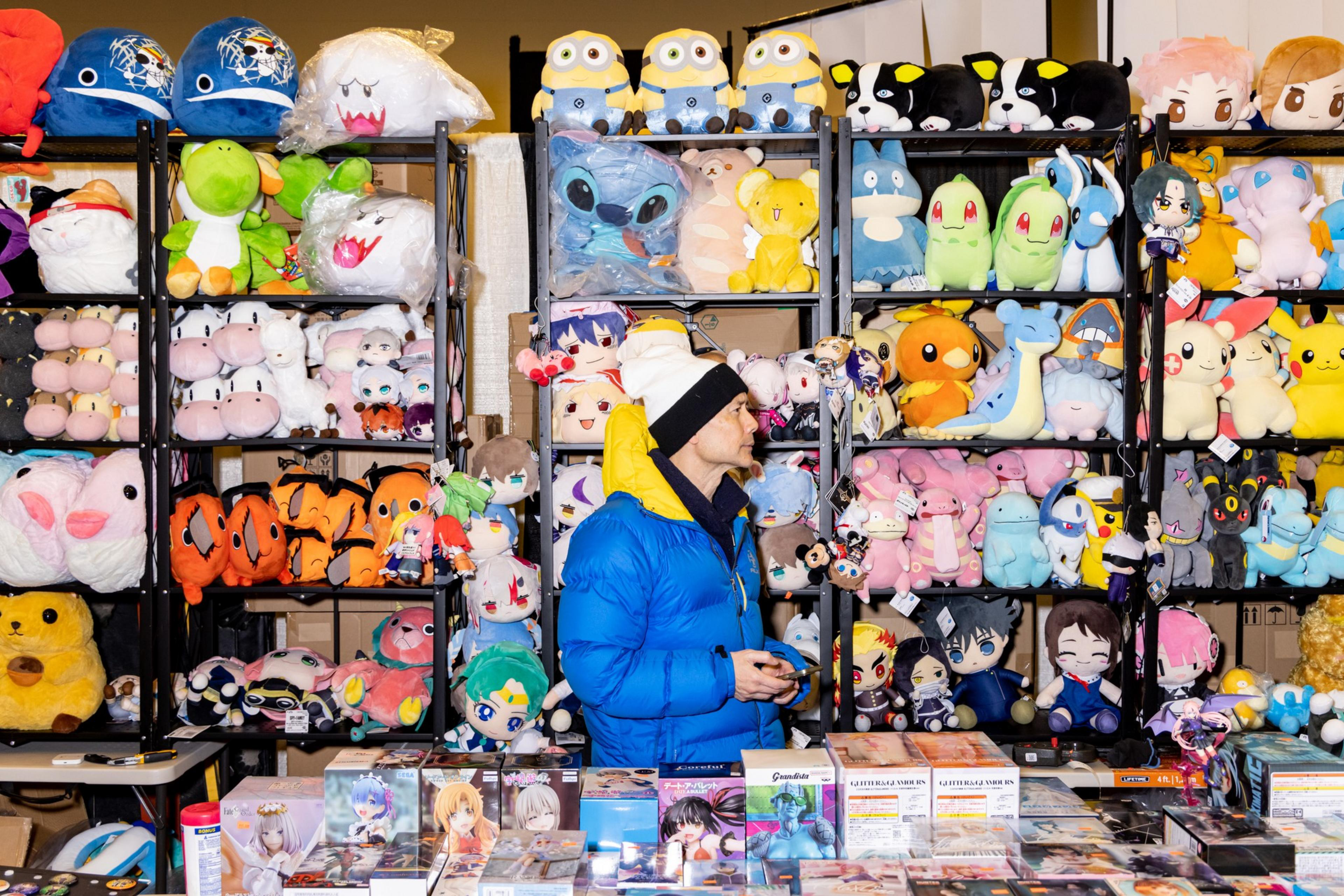 A person in a blue jacket and white beanie stands amid shelves of colorful plush toys, including characters like Pikachu and Stitch, at a convention booth.