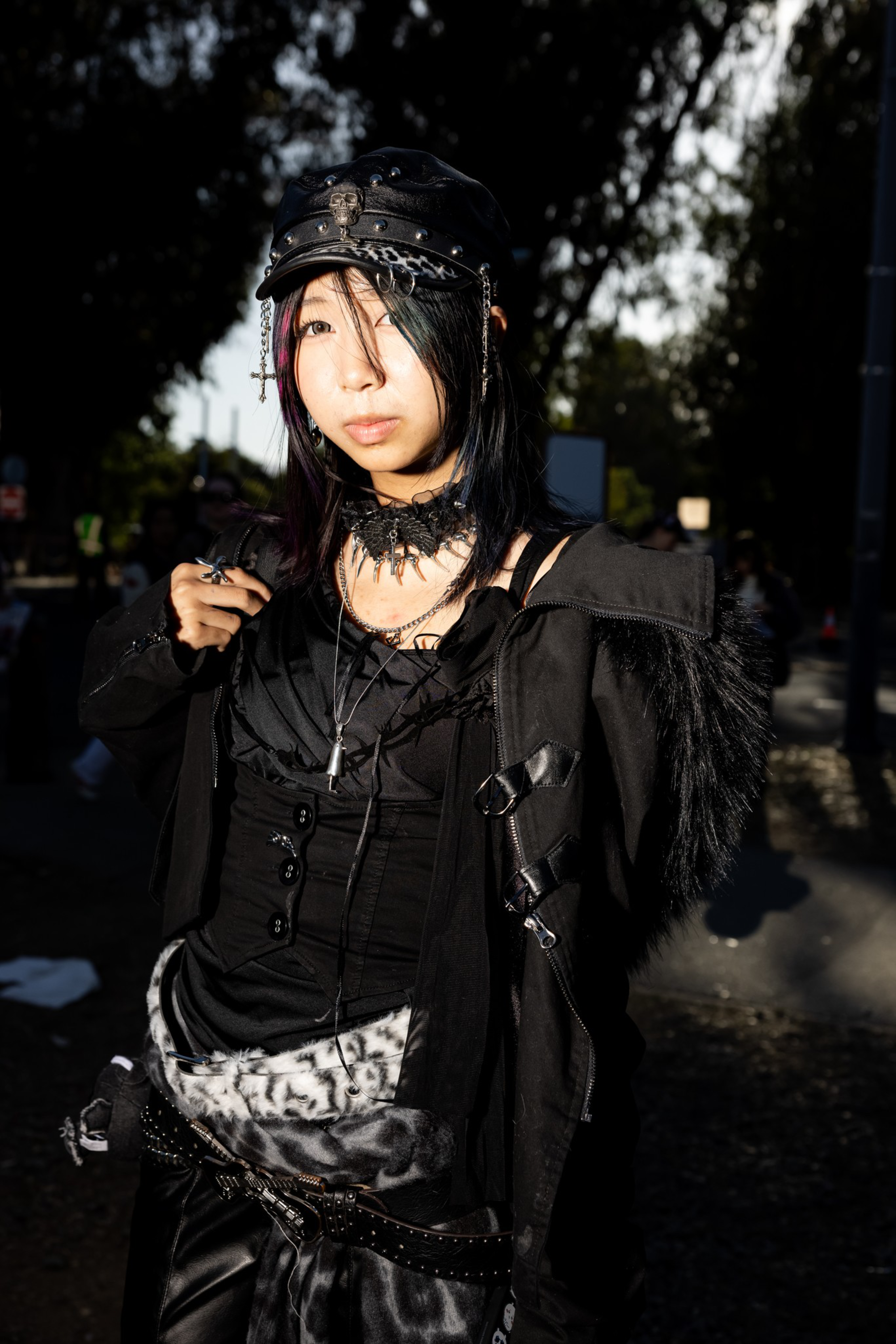A person with long black hair wears a black leather hat, spiked choker, layered black outfit with fur and metal accessories, standing outdoors in sunlight.