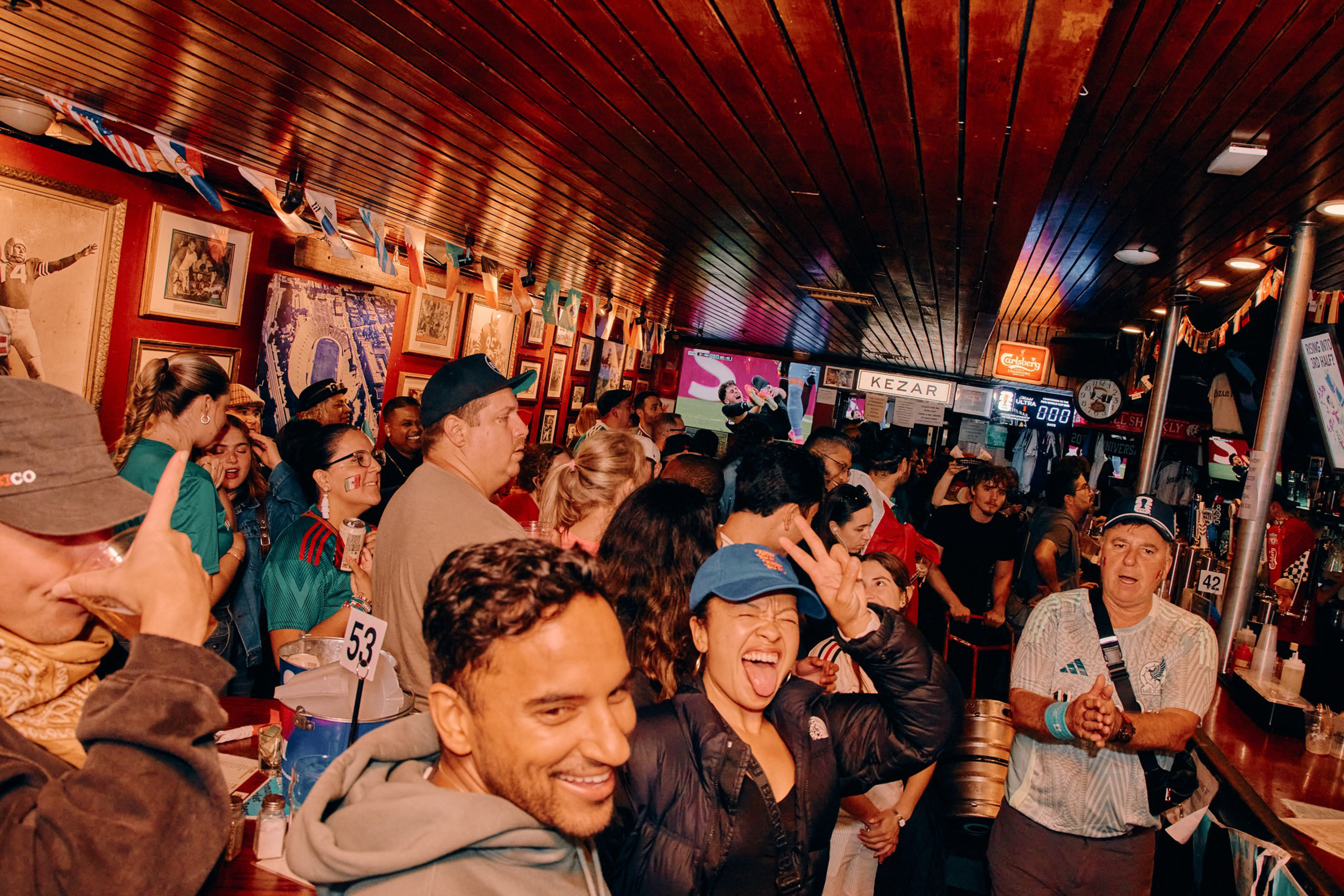 A lively bar crowded with people cheering and socializing, decorated with small flags and sports memorabilia, with two TVs showing a game.